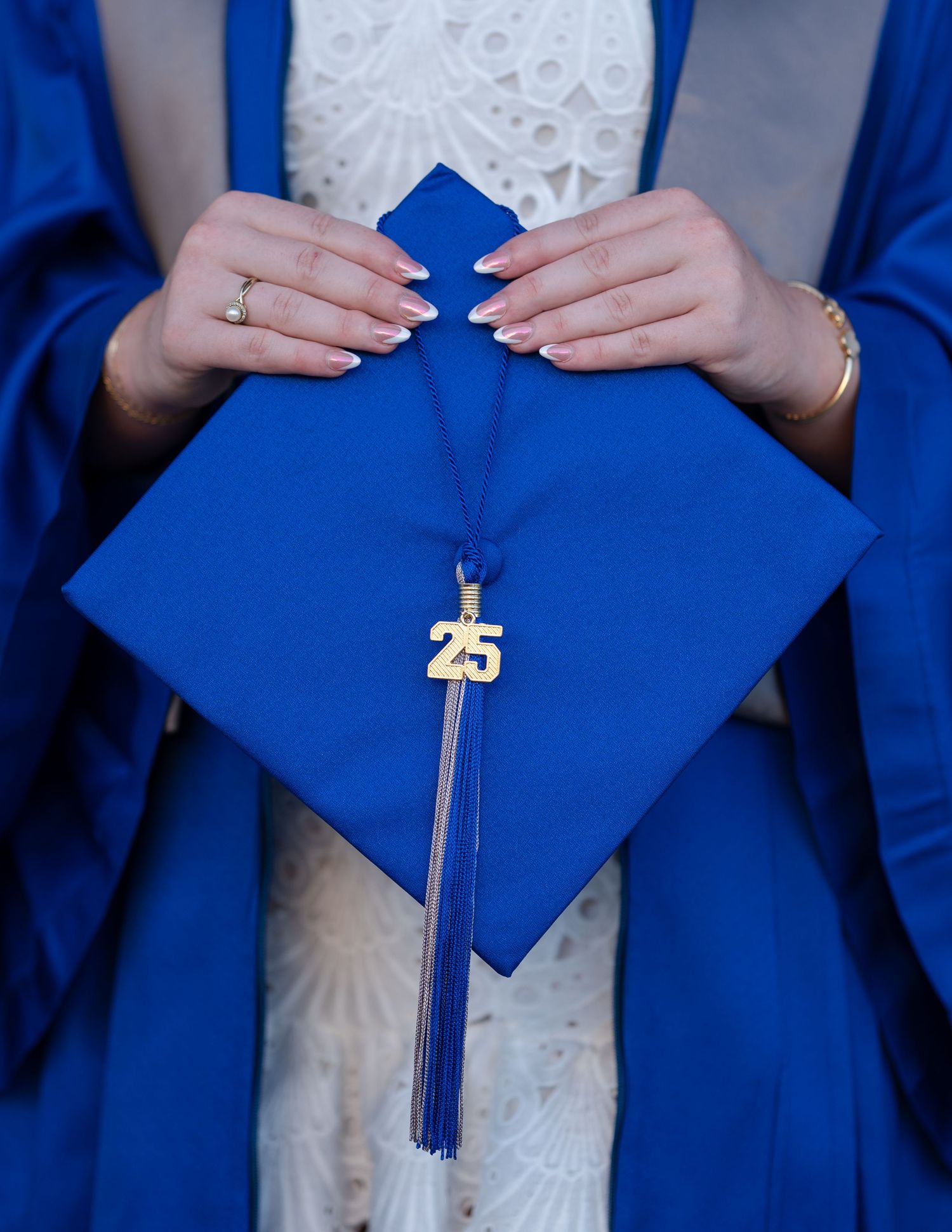 Hands hold a bright blue graduation cap with gold 25 tassel charm against a white lace dress and royal blue gown.
