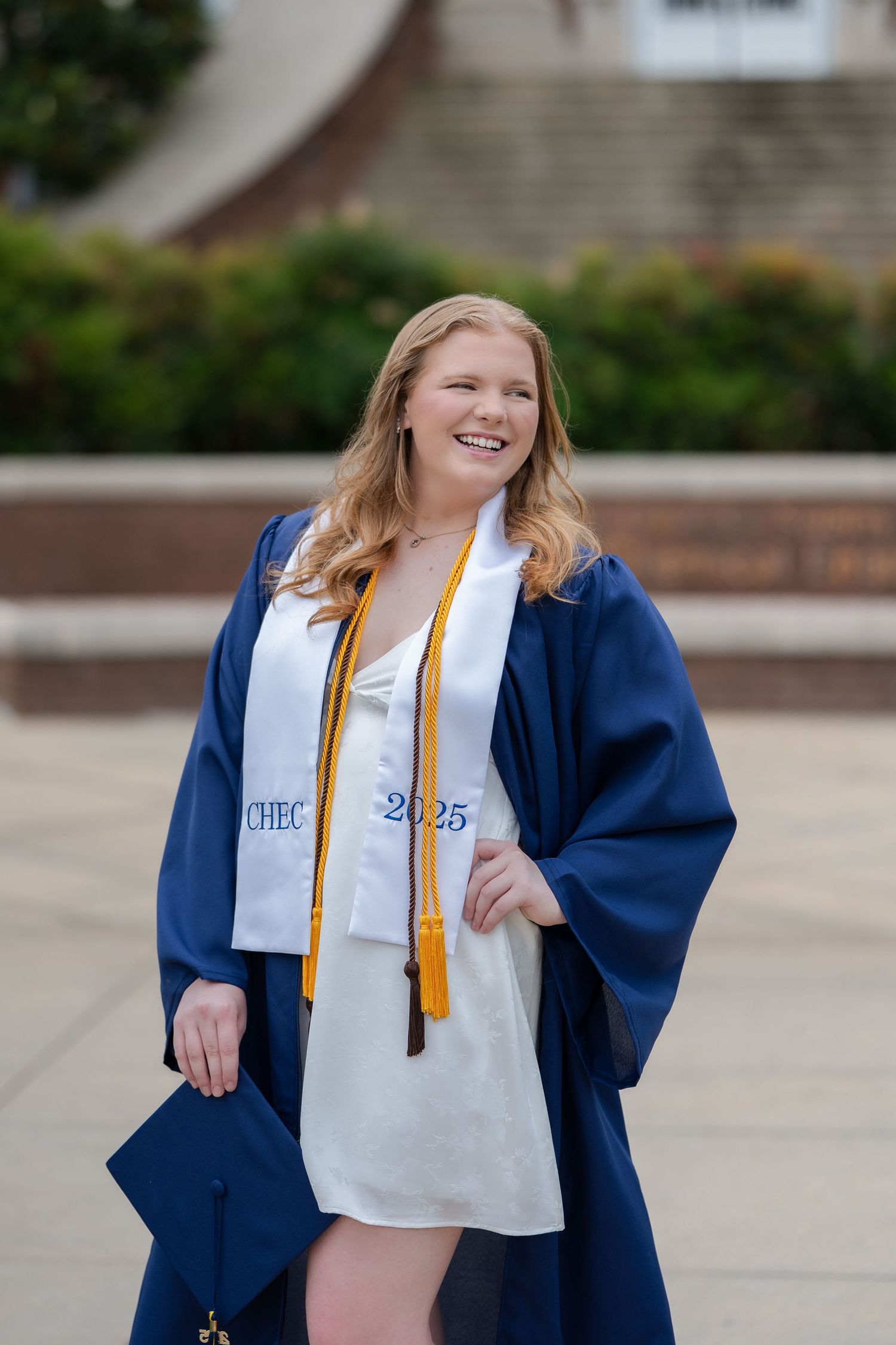 College graduate wearing a navy blue graduation gown, white dress and honor cords smiles outdoors during graduation.