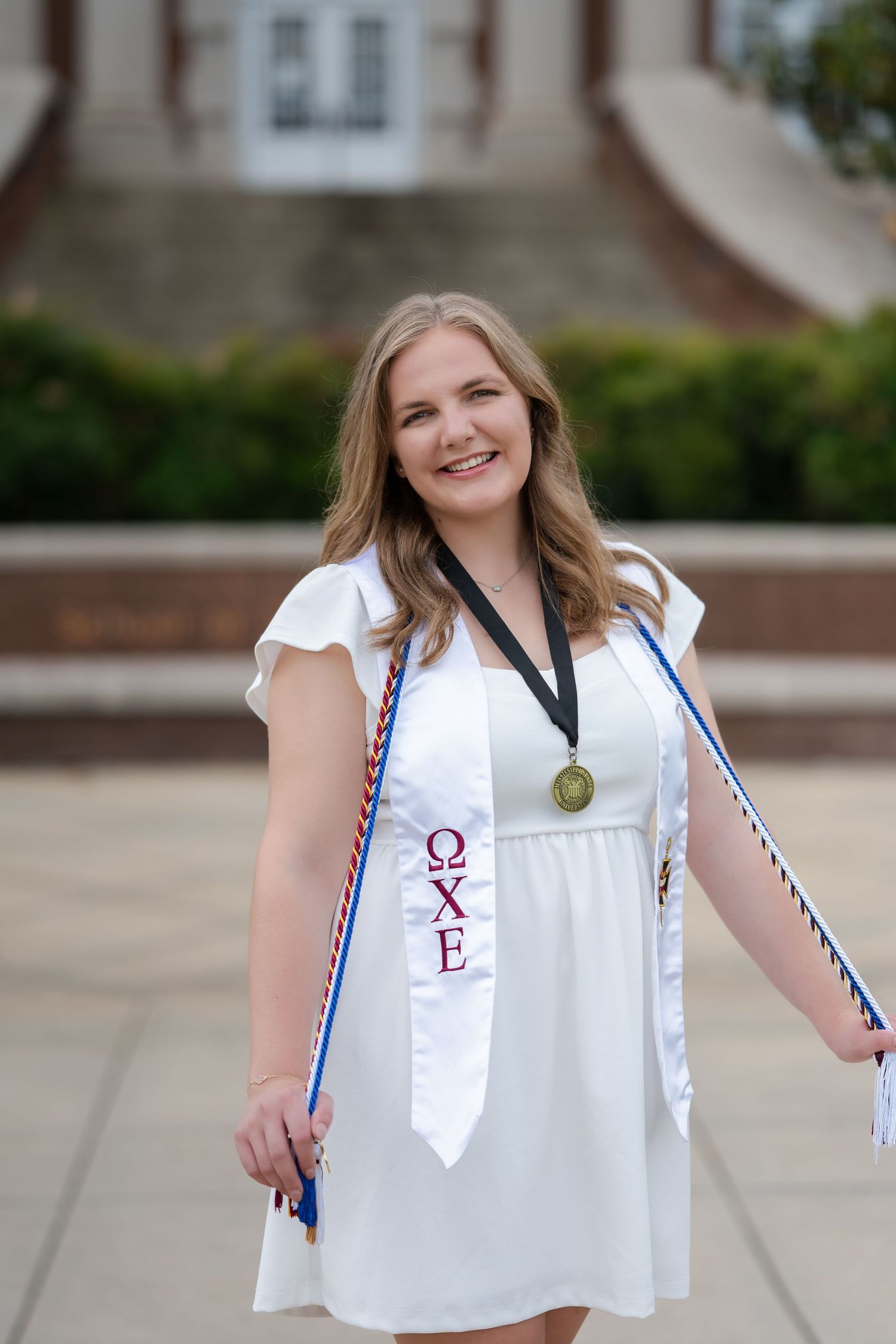 Graduate wearing white dress and graduation stole with sorority letters poses outdoors for graduation photo.