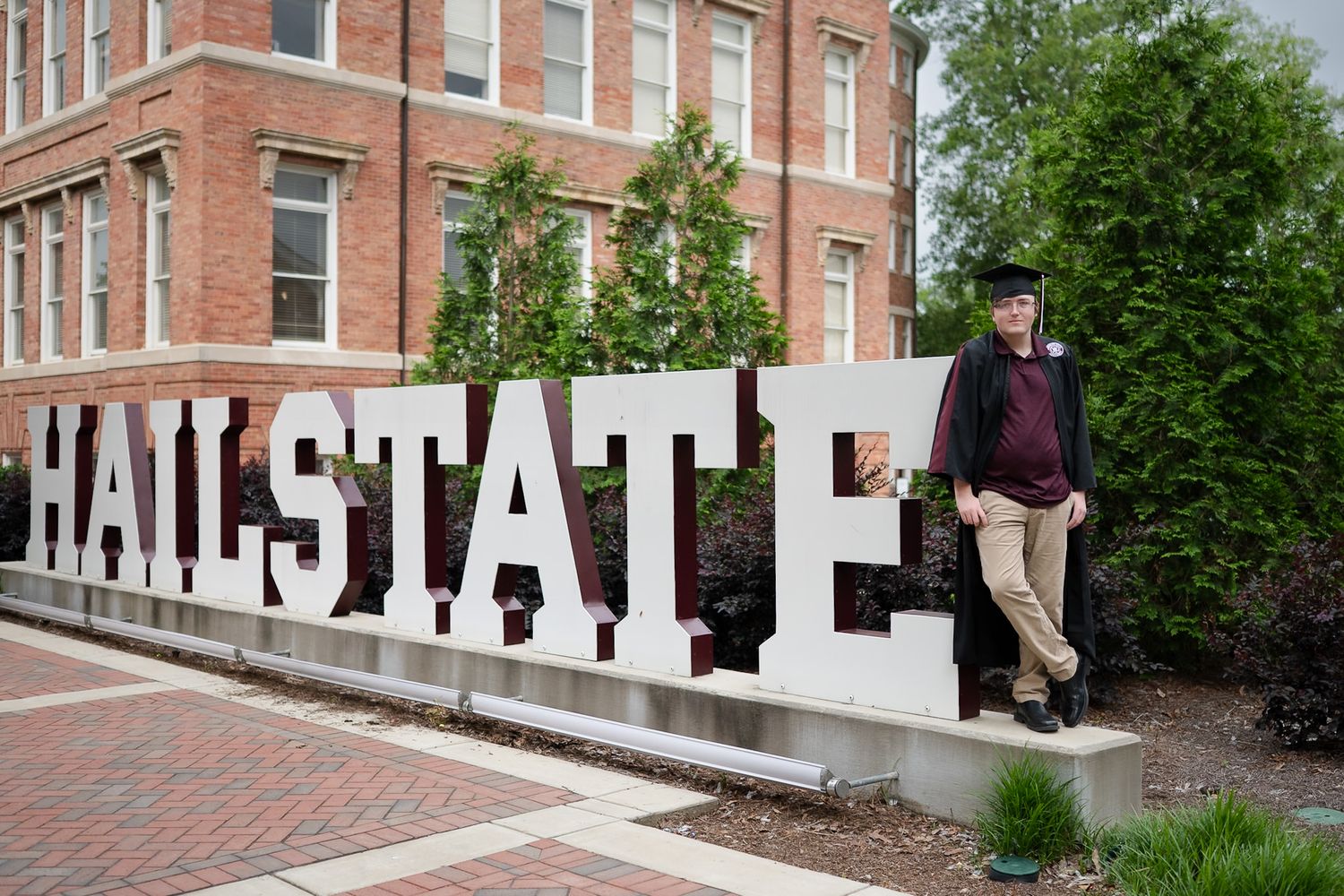A person stands near large white block letters spelling 'HAILSTATE' outside a brick university building on a spring day.
