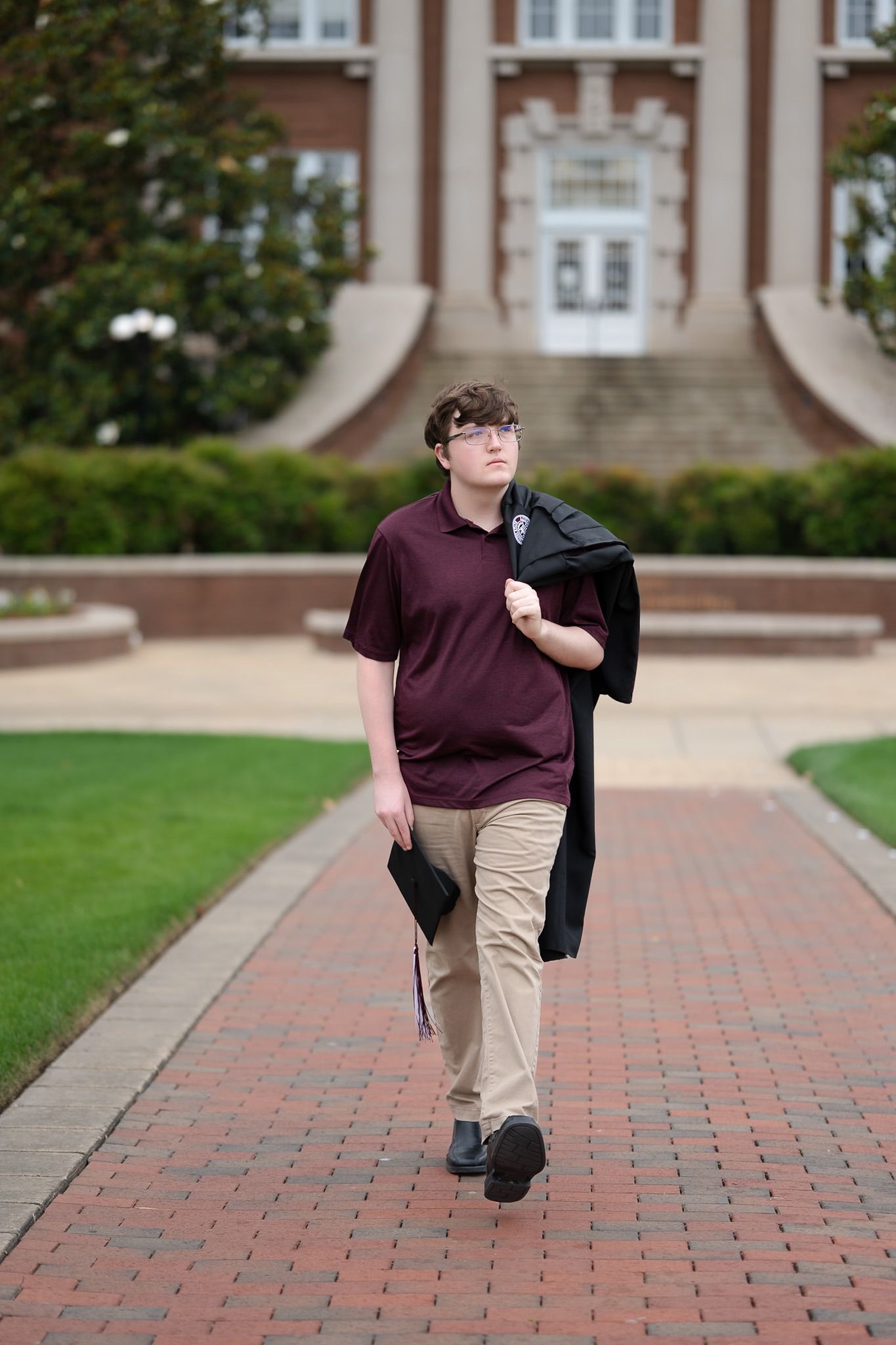 Student walking along brick pathway carrying graduation gown outside traditional brick university building.