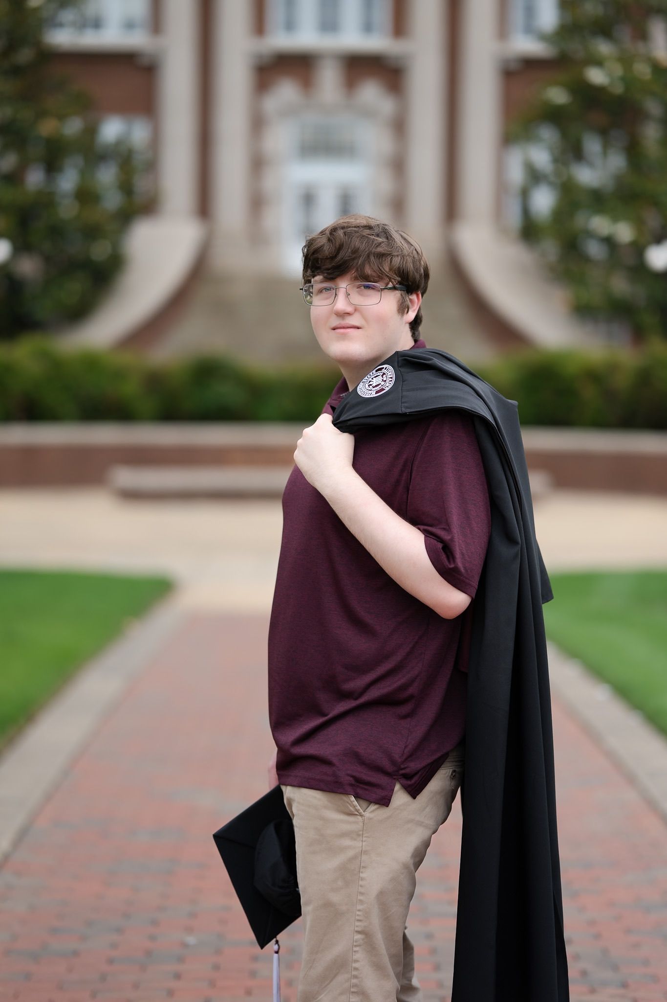 Student poses in a burgundy shirt and black graduation gown on a brick pathway in front of a collegiate building.