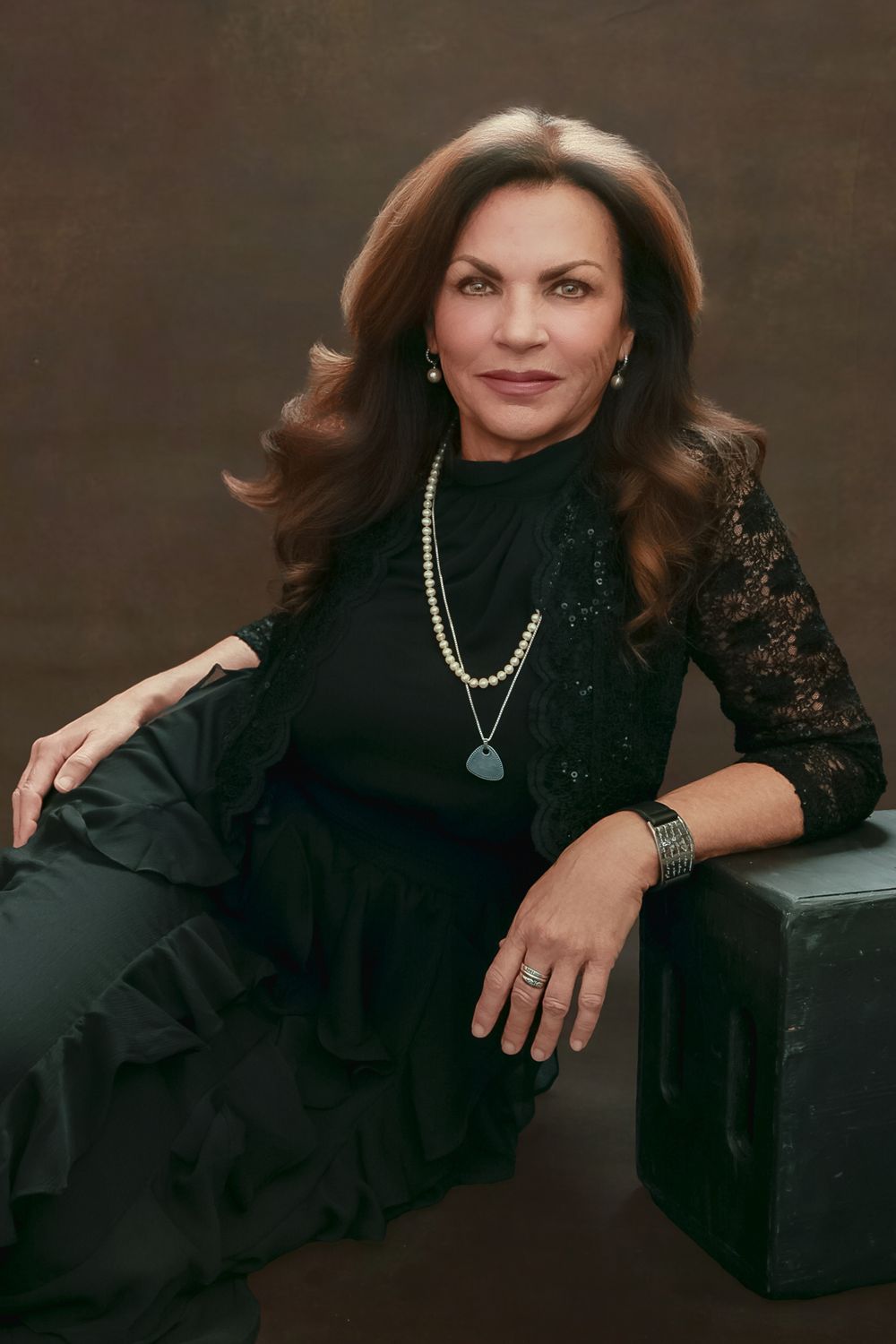 Studio portrait of someone in a black lace dress and pearl necklace sitting elegantly against a dark background.