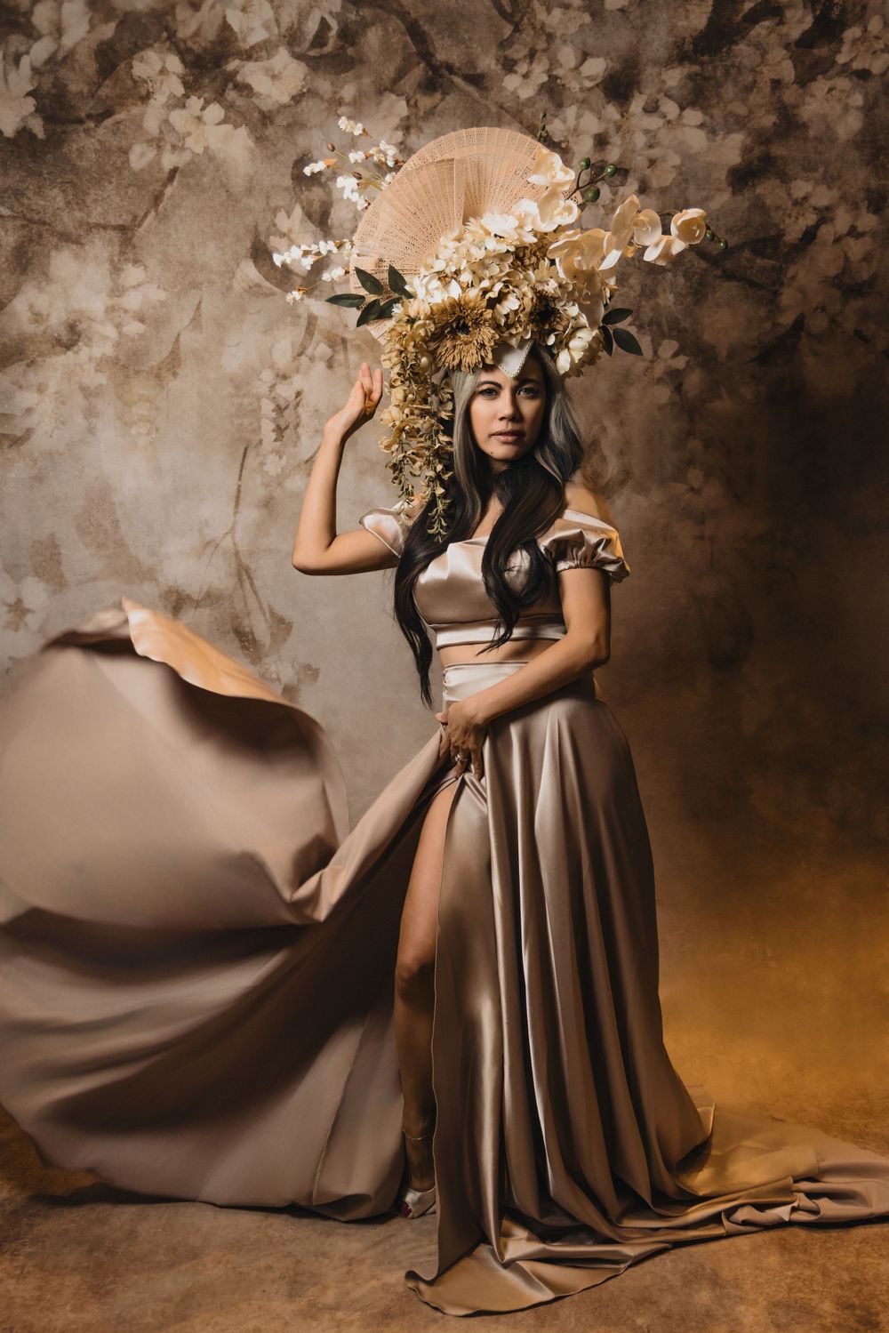 A flowing mauve dress billows dramatically in a moody brown studio setting with delicate floral elements.