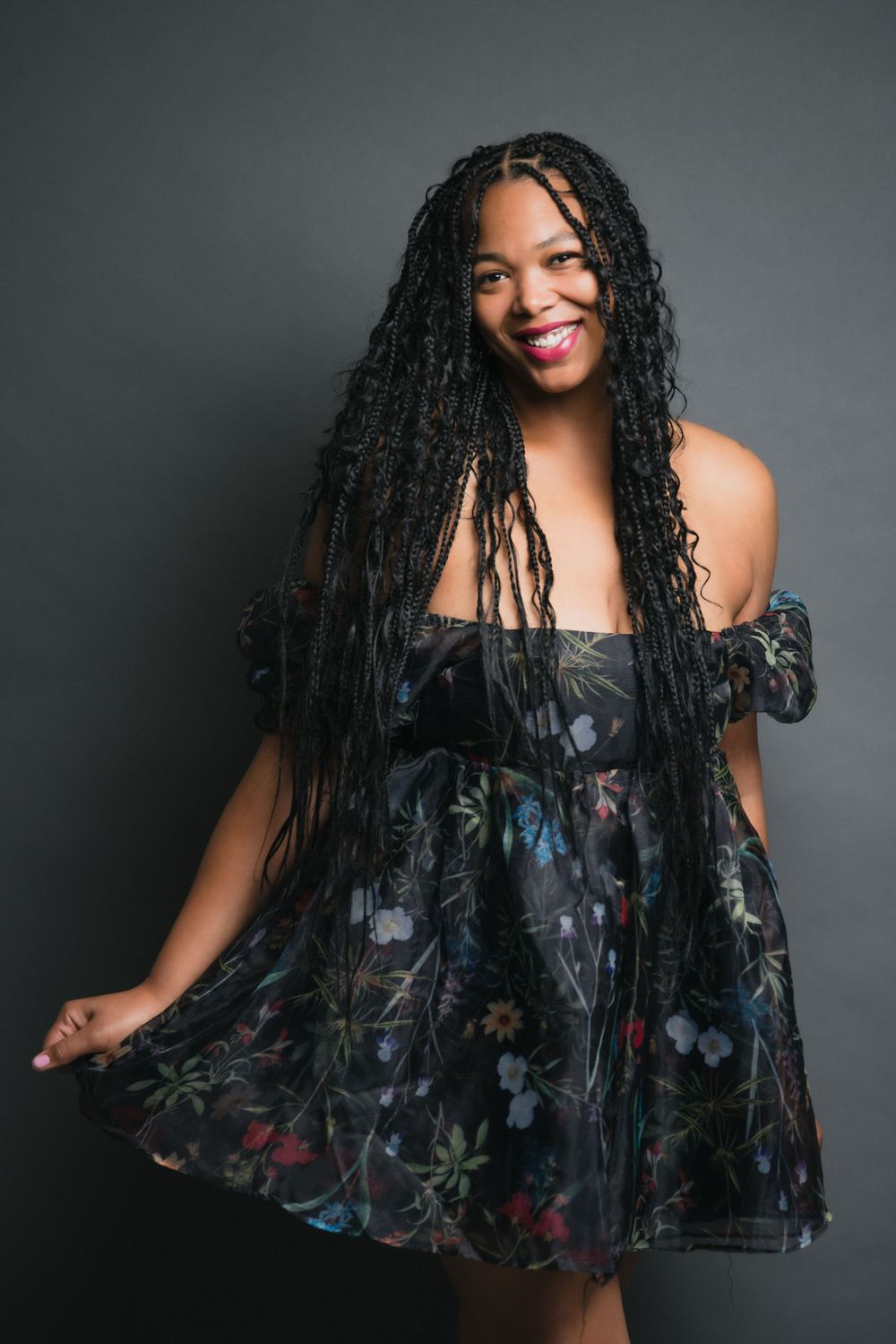 Smiling woman in off-shoulder floral dress with long braided hair poses against dark background.