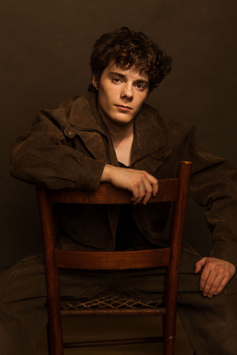 Moody artistic portrait of someone in brown jacket sitting backwards on wooden chair against dark background.