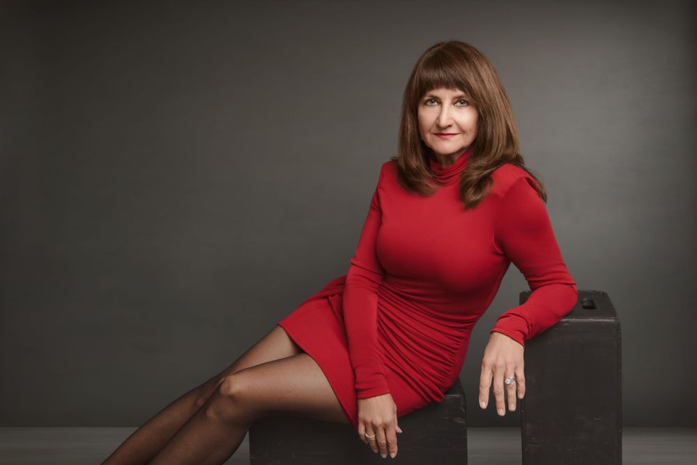 A person in a fitted red dress with long sleeves poses elegantly on a dark chair against a grey backdrop.