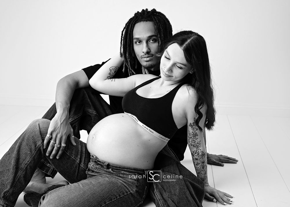 Black and white maternity photo of a pregnant woman and her partner sitting together on a studio floor.