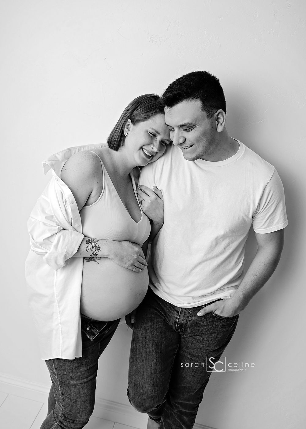Black and white maternity photo of a smiling pregnant woman and man leaning together affectionately in a studio setting.