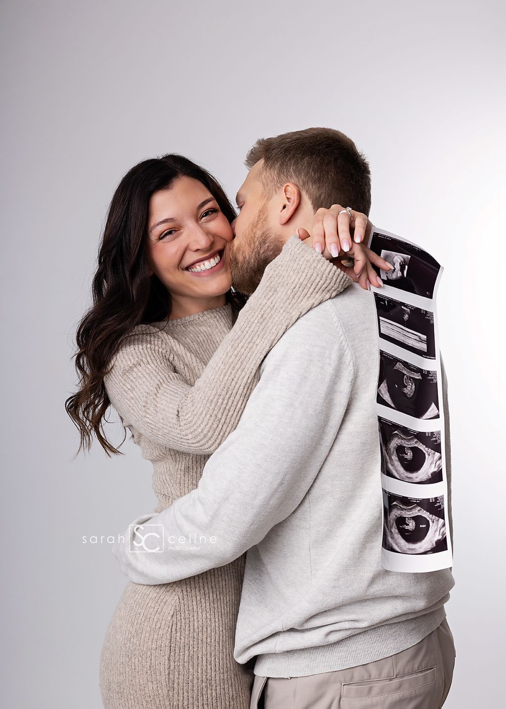 Couple in neutral sweaters embraces while holding ultrasound photos, sharing a joyful pregnancy announcement moment.