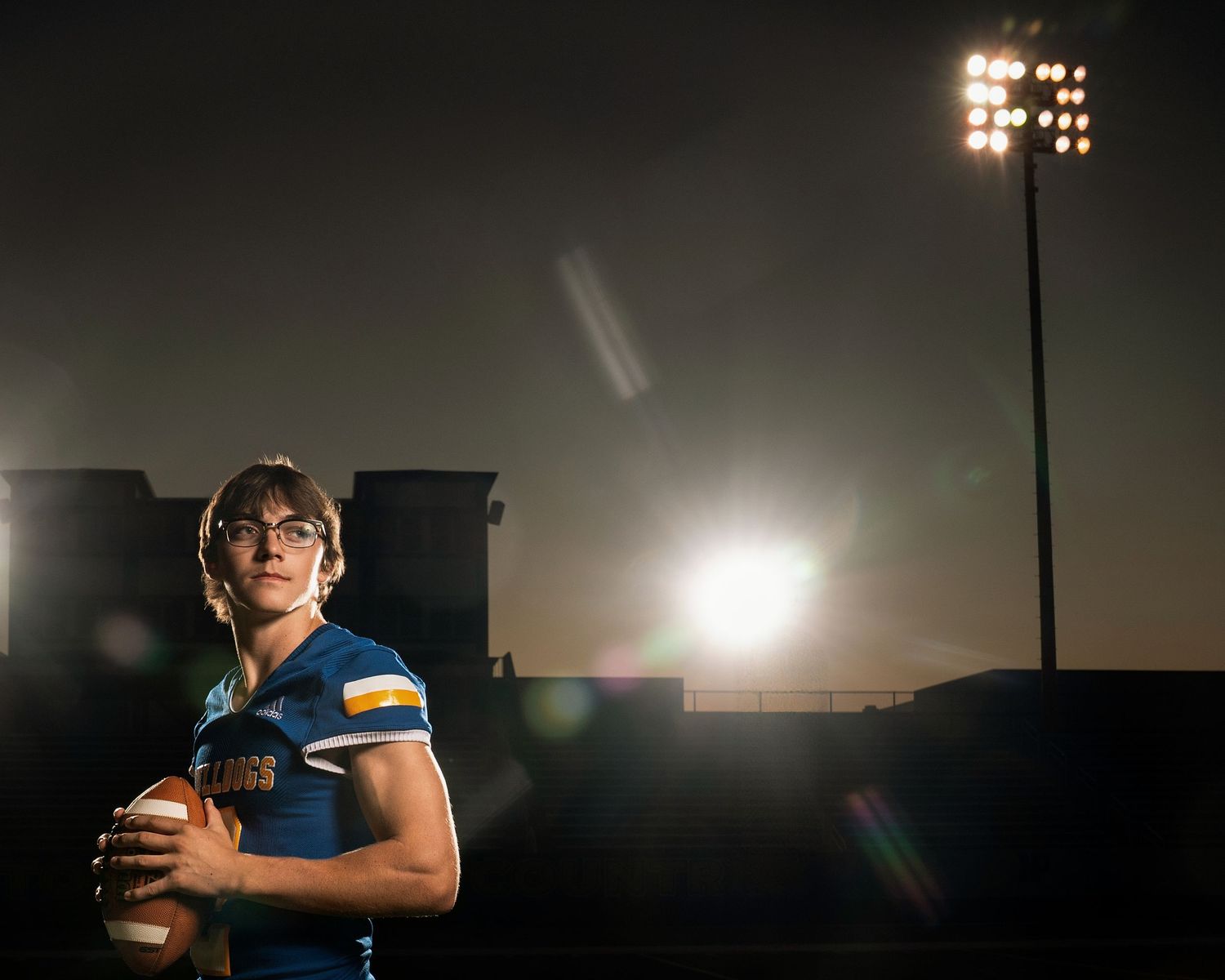 A silhouetted figure stands in a dimly lit stadium under floodlights creating a dramatic atmospheric scene.