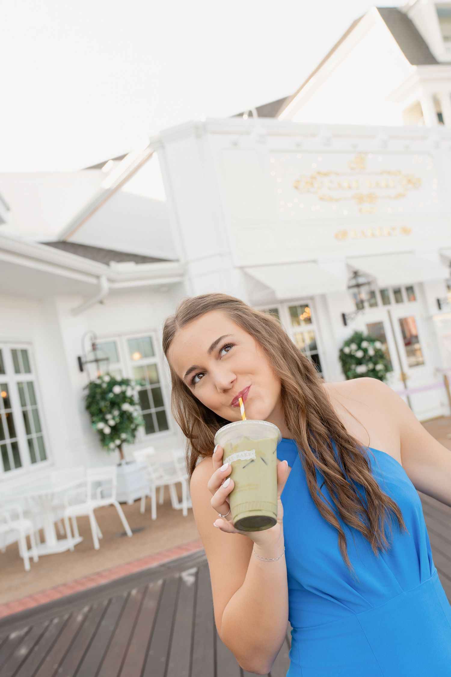 Person in bright blue top enjoying a beverage on a white porch with greenery and windows visible in background.