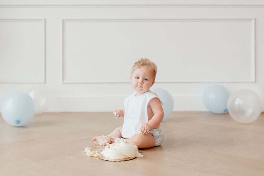 Baby boy in white onesie sits on floor smashing first birthday cake surrounded by blue balloons.