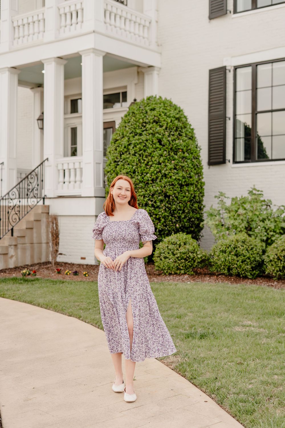 Red-haired woman in floral midi dress standing on pathway before elegant white mansion with columns.