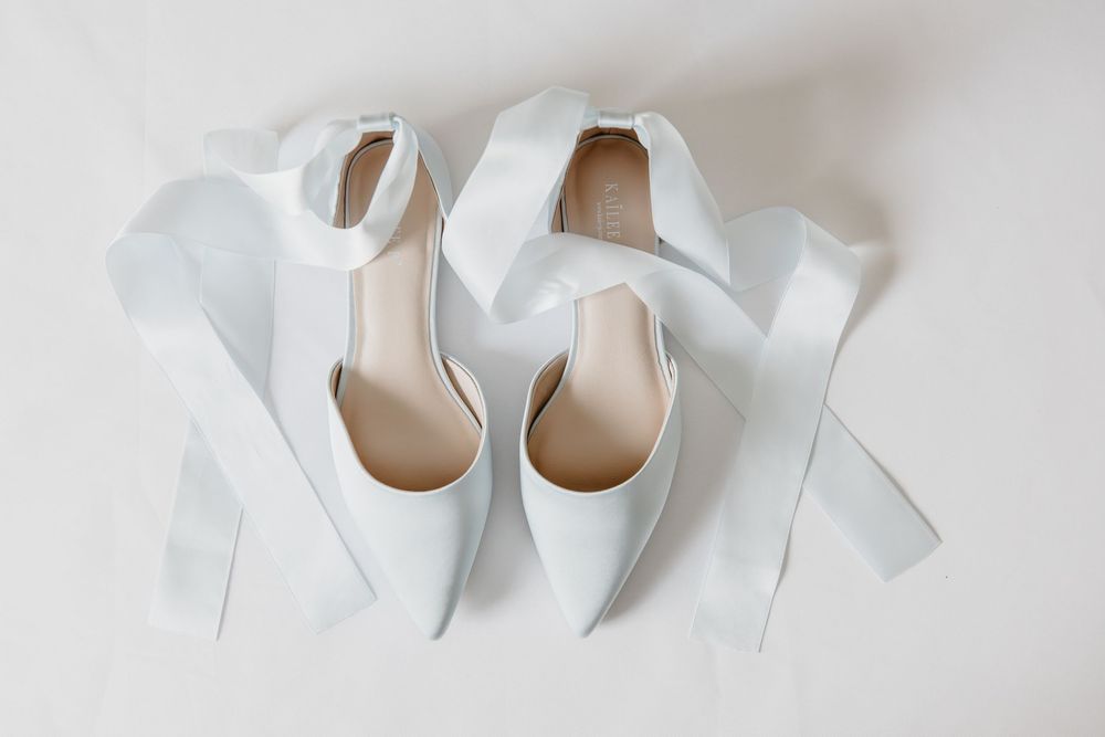 White pointed-toe bridal heels with elegant satin ribbon ankle ties displayed on a light background.