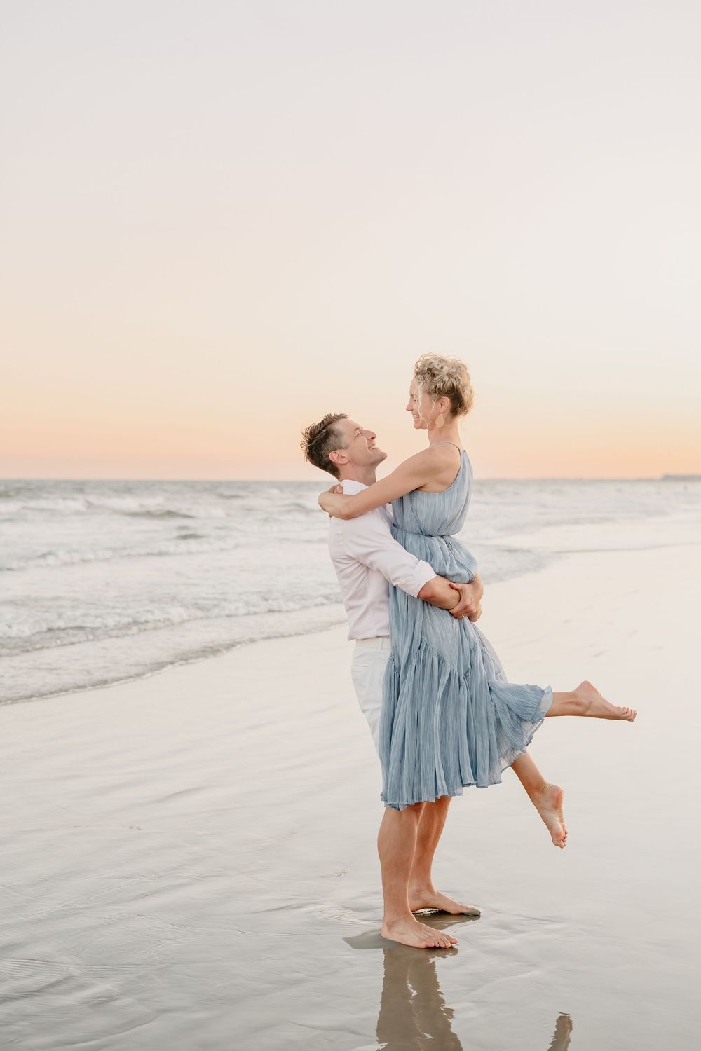 A romantic couple embraces on a beach at sunset, with soft waves and pastel sky in the background.