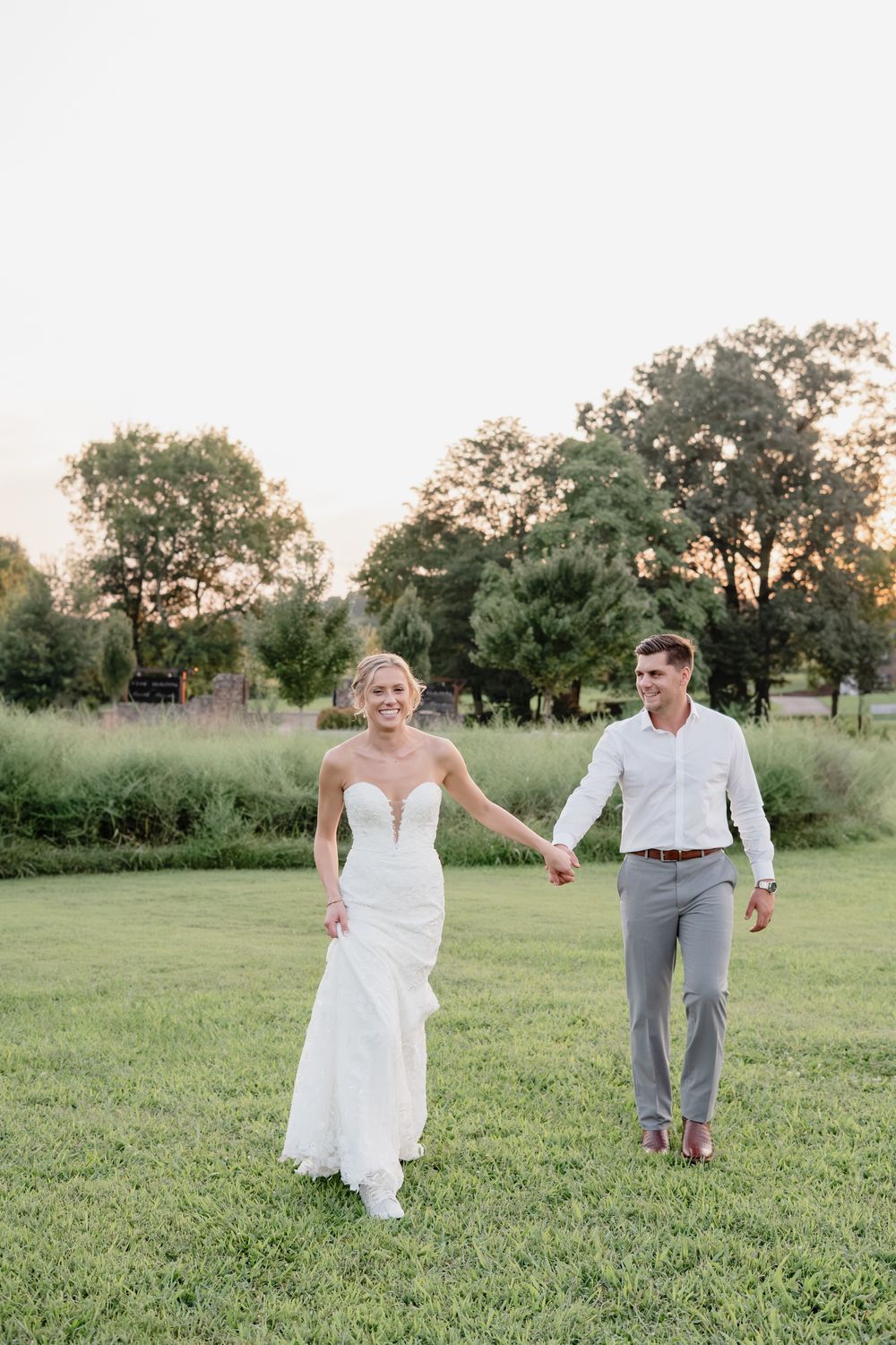 Smiling bride in white gown holds grooms hand walking across green lawn at golden hour outdoor wedding.