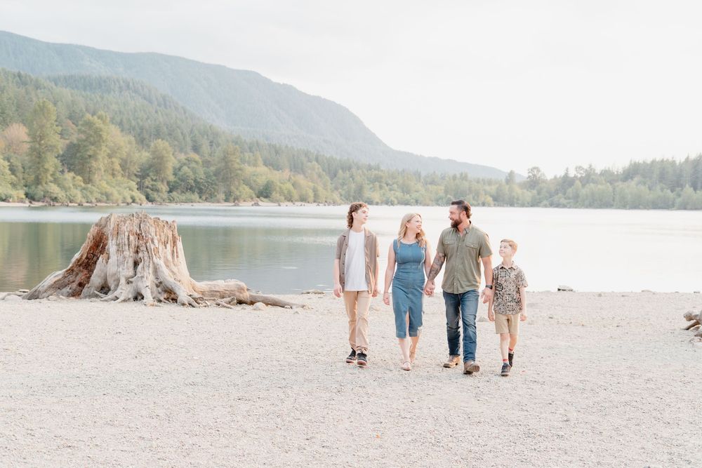 Family of four walking hand-in-hand on a sandy beach beside a calm lake surrounded by lush forest and mountains.