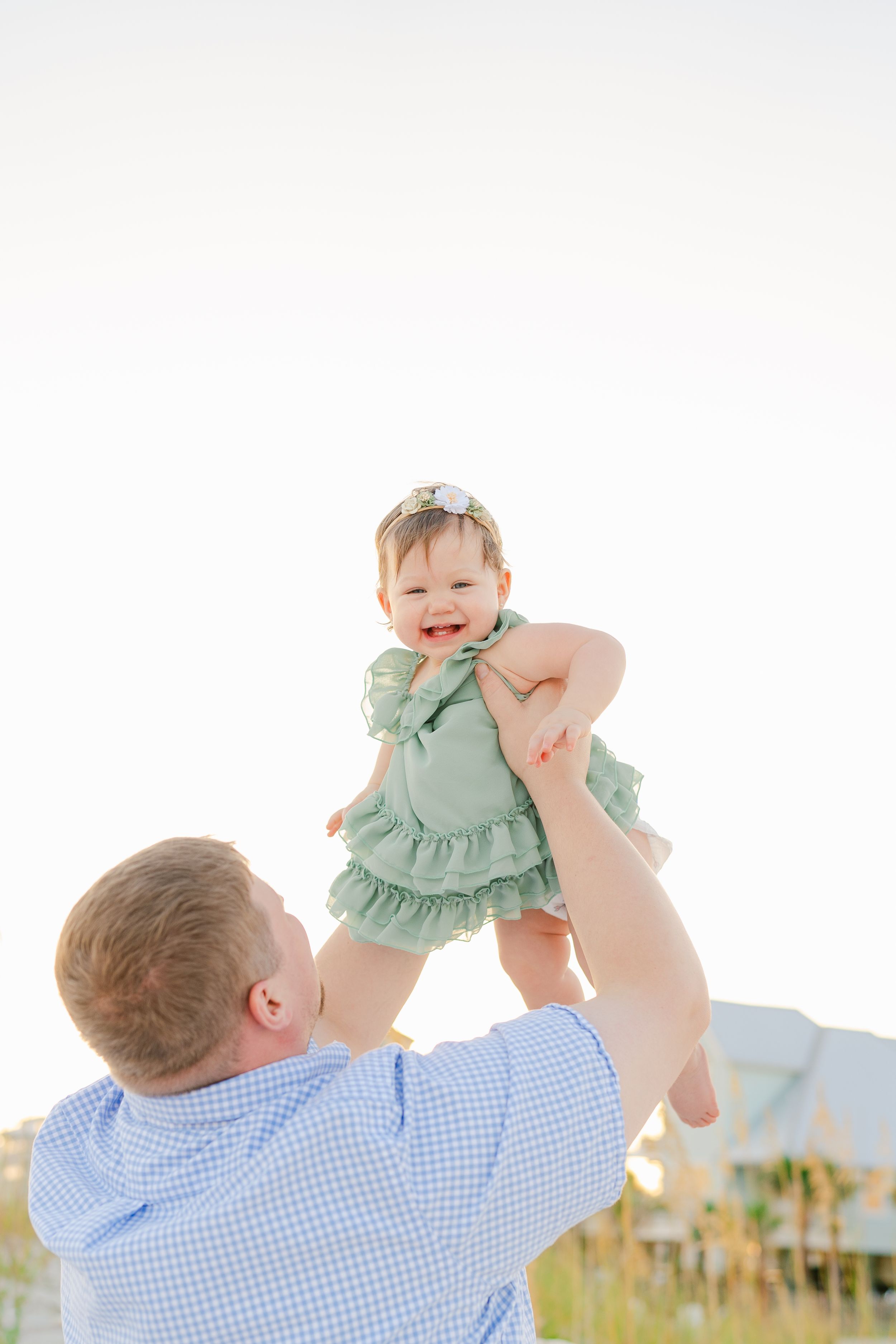 Parent lifts joyful baby wearing mint green outfit high in the air against bright sky.
