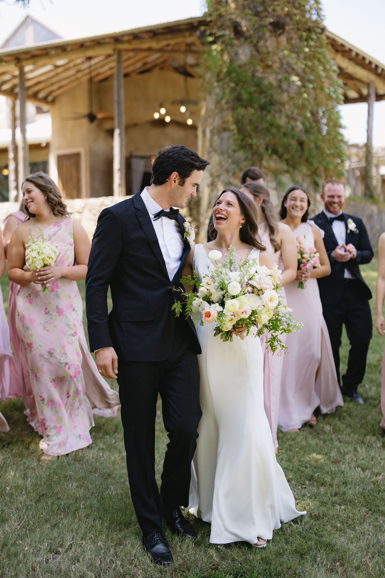 Laughing bride and groom walk together outdoors followed by bridesmaids in pink dresses after wedding ceremony.