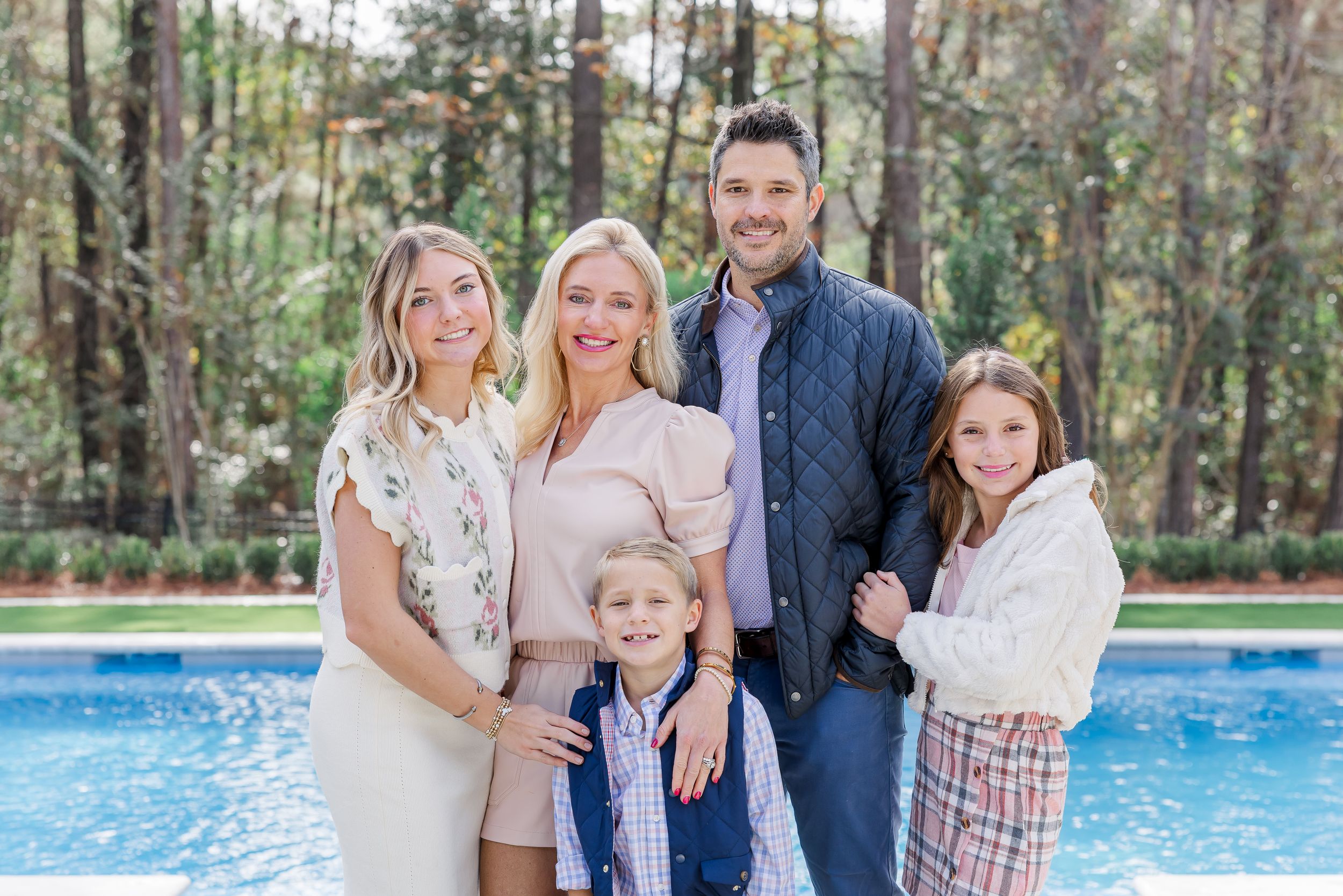 A happy family poses together outdoors near a swimming pool on a sunny day surrounded by tall pine trees.