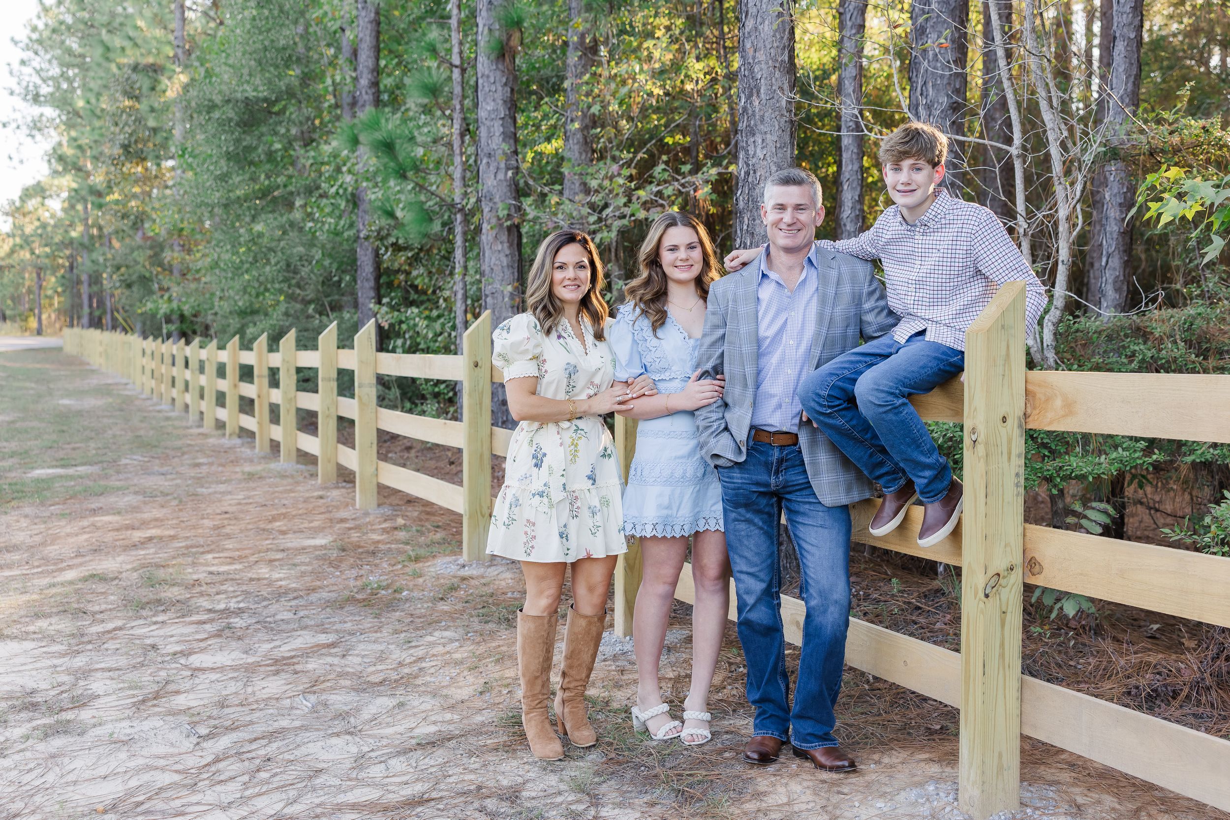 A family poses together on a wooden fence walkway surrounded by trees during an outdoor photo session.