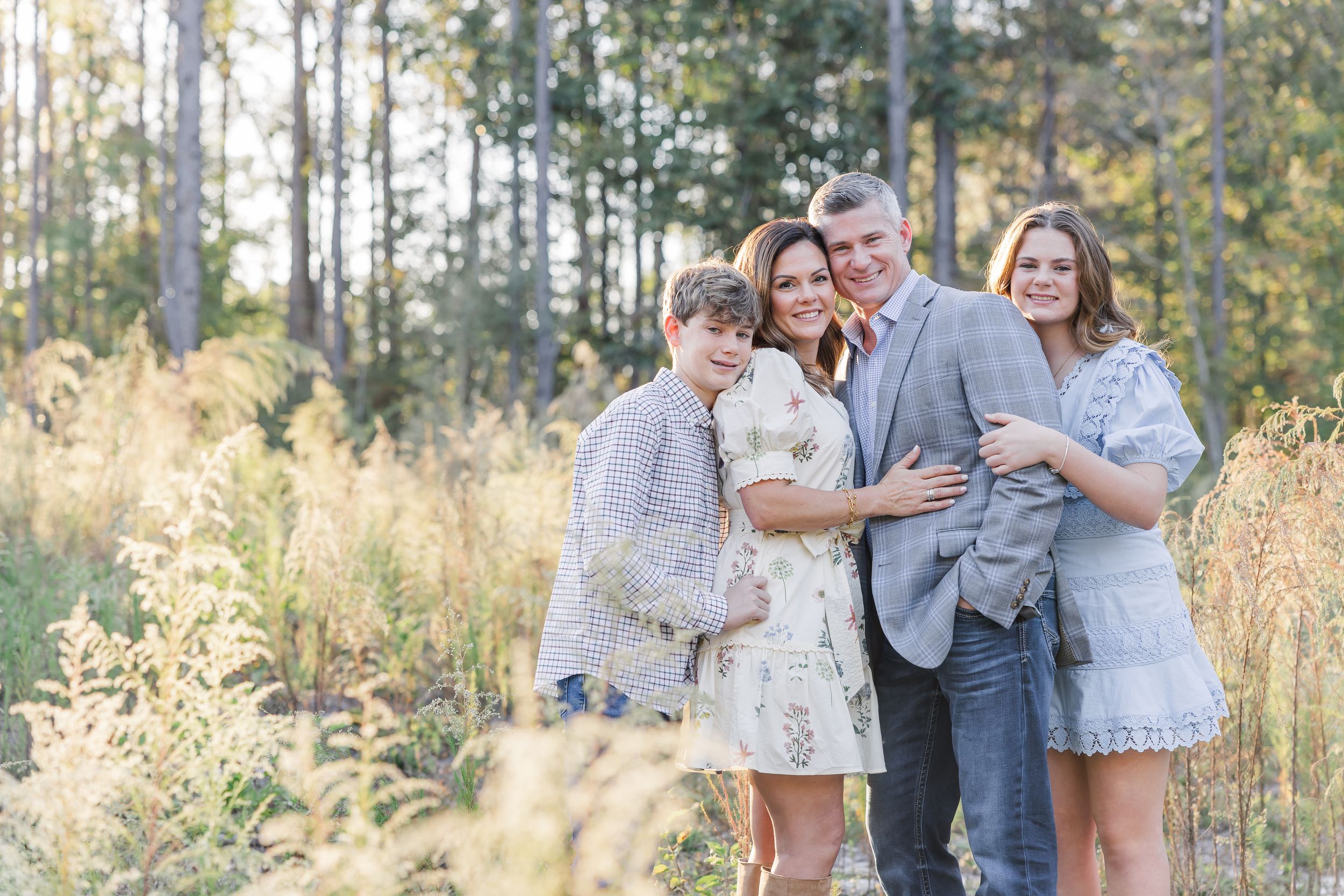 A group poses together outdoors in a sunlit field with tall grass and pine trees in the background.