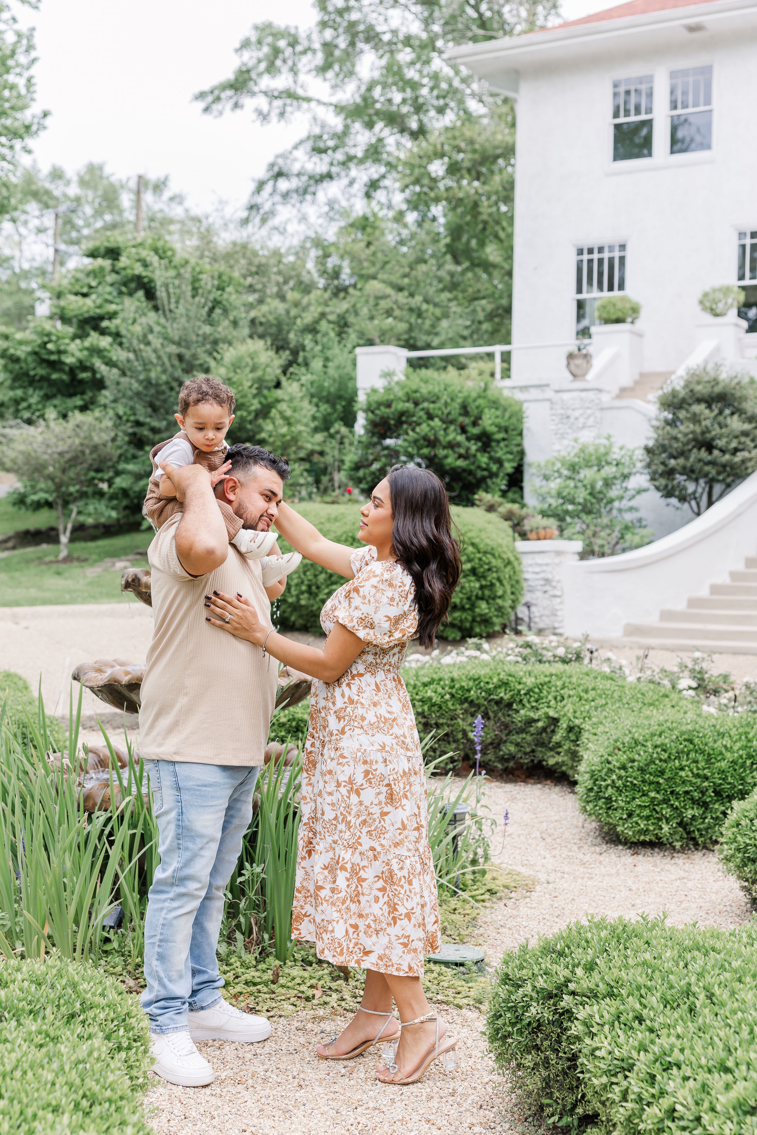 A couple shares a tender dance moment together in a lush garden setting with a white building in the background.