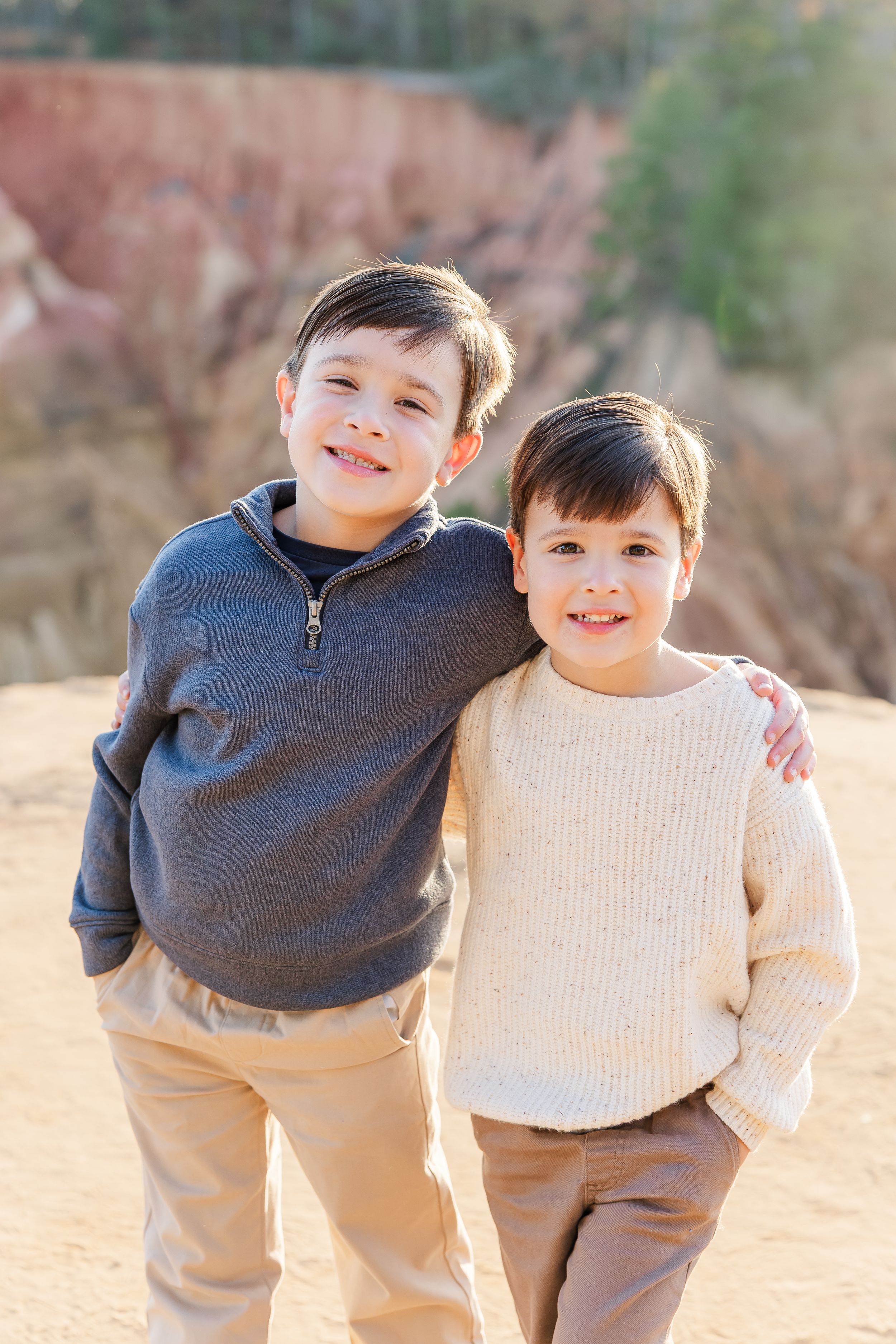 Two young children in coordinating sweaters and khaki pants pose together outdoors on a sunny autumn day.