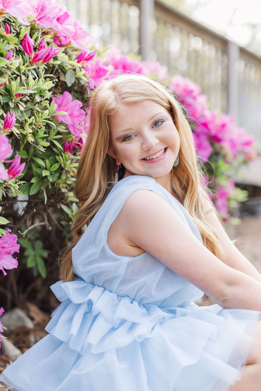 A light blue ruffled dress is worn while posing near pink azalea flowers and a white fence in a spring garden setting.