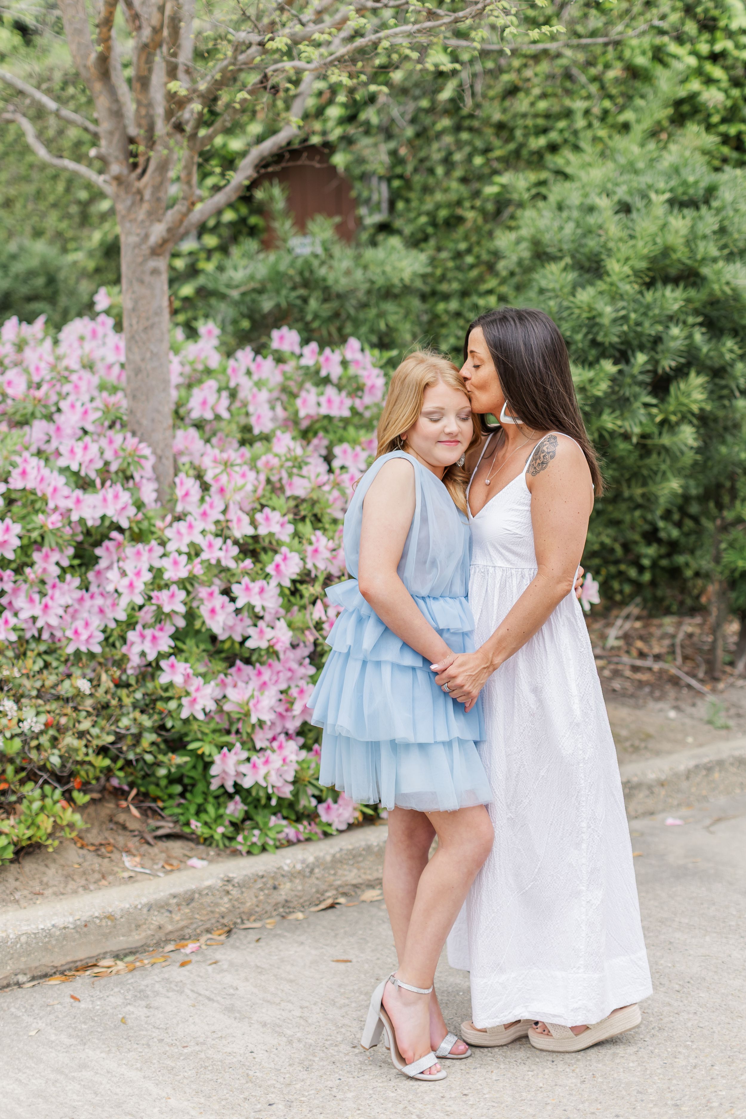A mother and daughter share tender moments in a garden with blooming pink azaleas and trees in the background.
