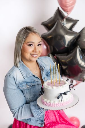 Person in denim jacket poses with pink birthday cake and black balloons for celebratory photo shoot.