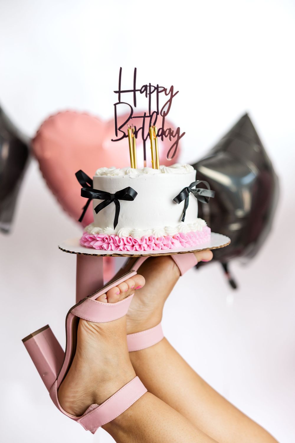 A white birthday cake with pink trim and black bows held up by feet wearing pink block heel sandals.