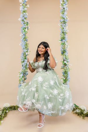 A young child in a mint green floral dress poses playfully in front of a floral backdrop with hanging vines.