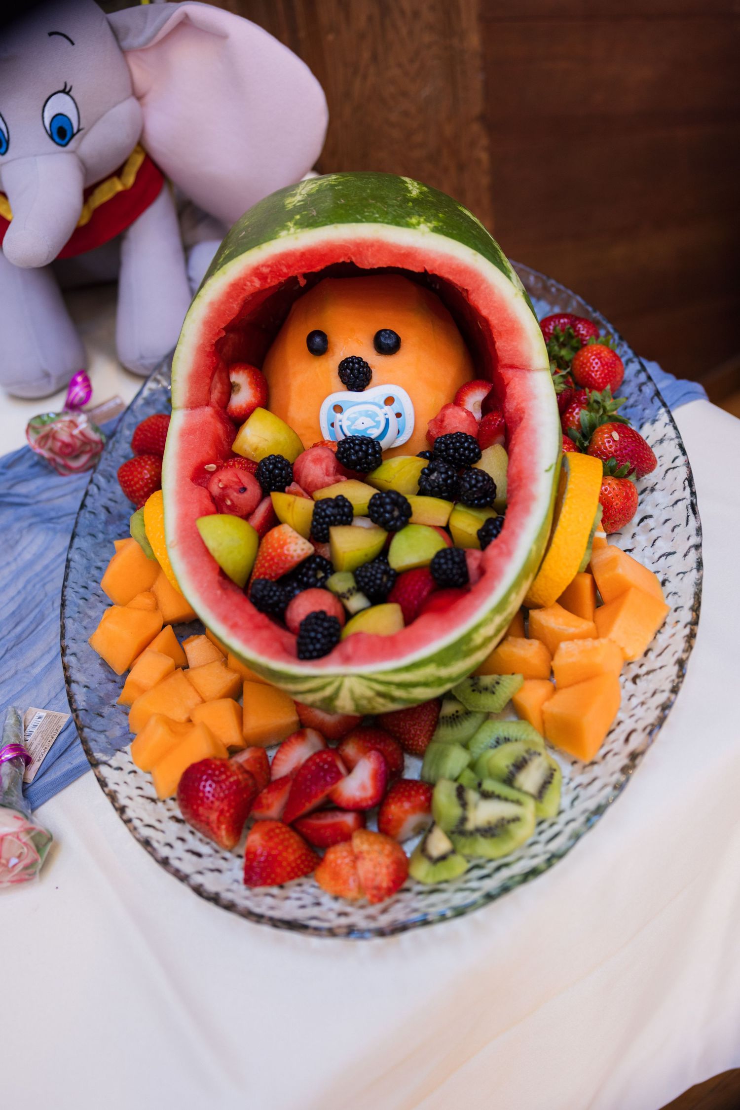 Creative baby shower watermelon carving shaped like a baby in a cradle, surrounded by fresh fruit on a platter.