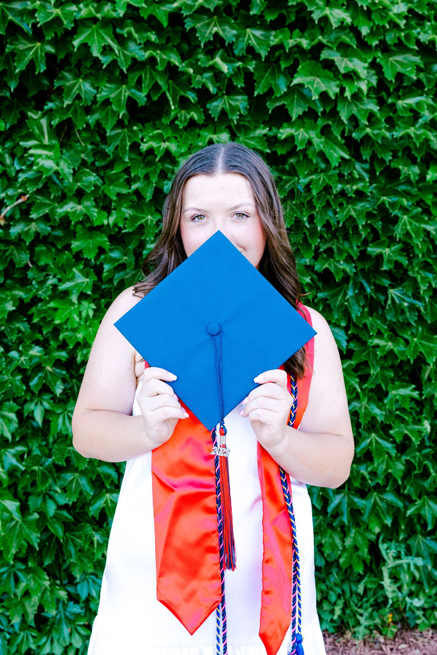 Graduate holding a blue graduation cap in front of a green ivy wall wearing red and white regalia.