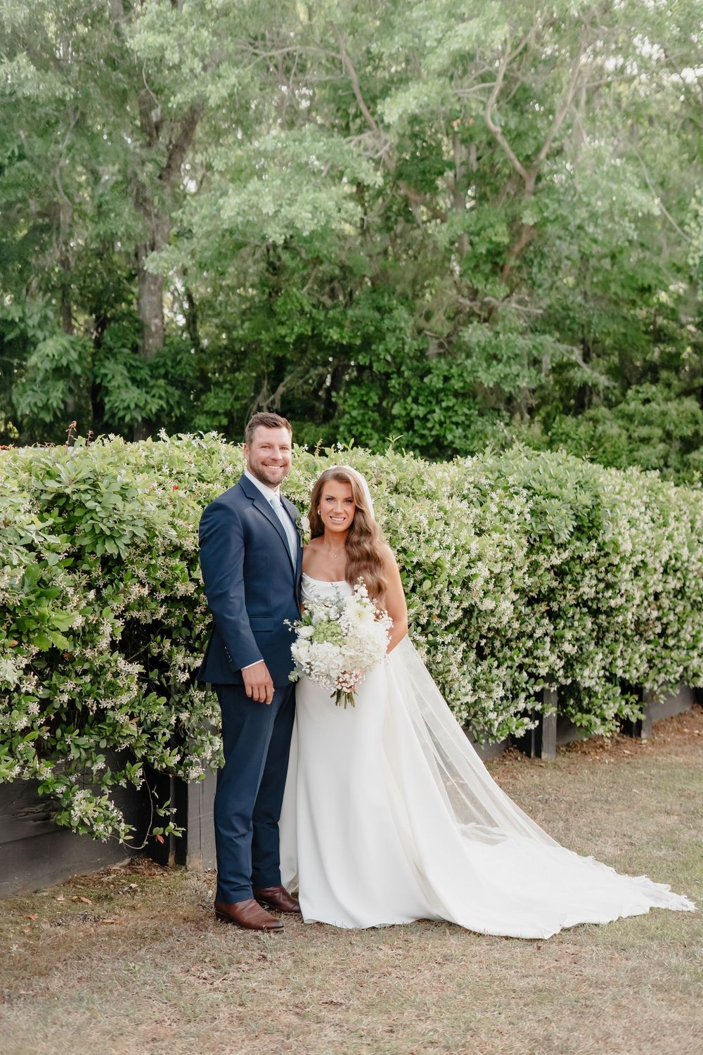 A newlywed couple stands together outdoors in front of lush green hedges, with the bride in white and groom in navy.
