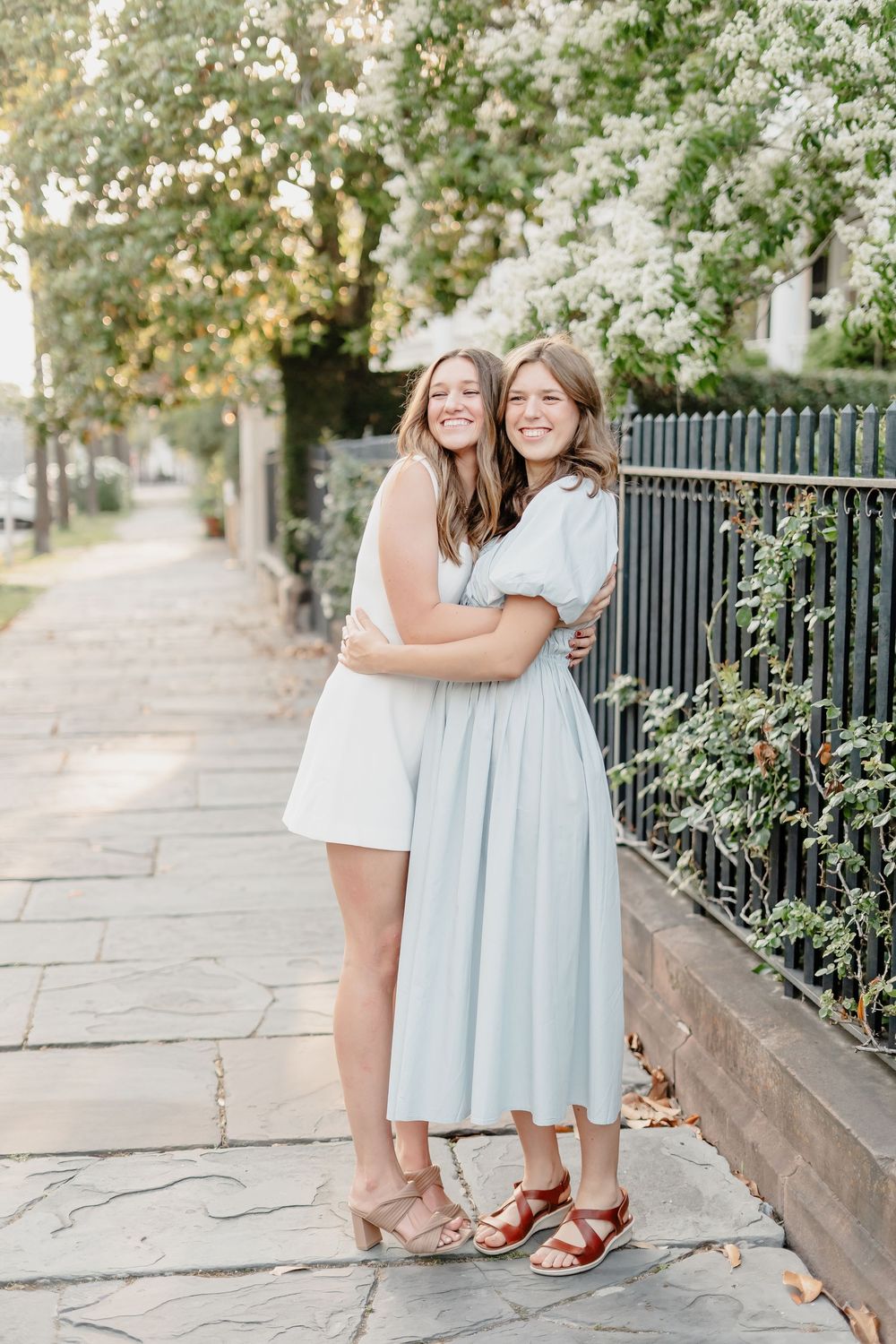 Two people in pastel dresses share an outdoor moment together on a tree-lined sidewalk.