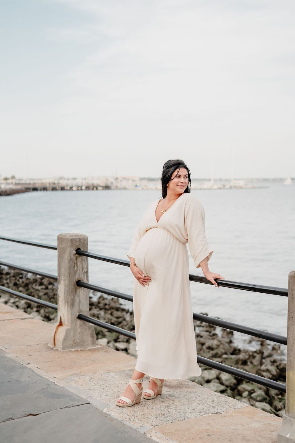 Expectant mother in flowing cream maxi dress poses along waterfront railing during sunset maternity photoshoot.
