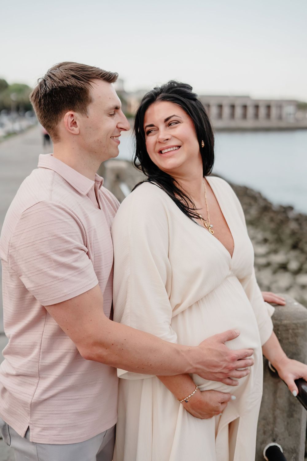 Couple shares romantic moments by the waterfront in matching light pink and cream outfits during sunset.