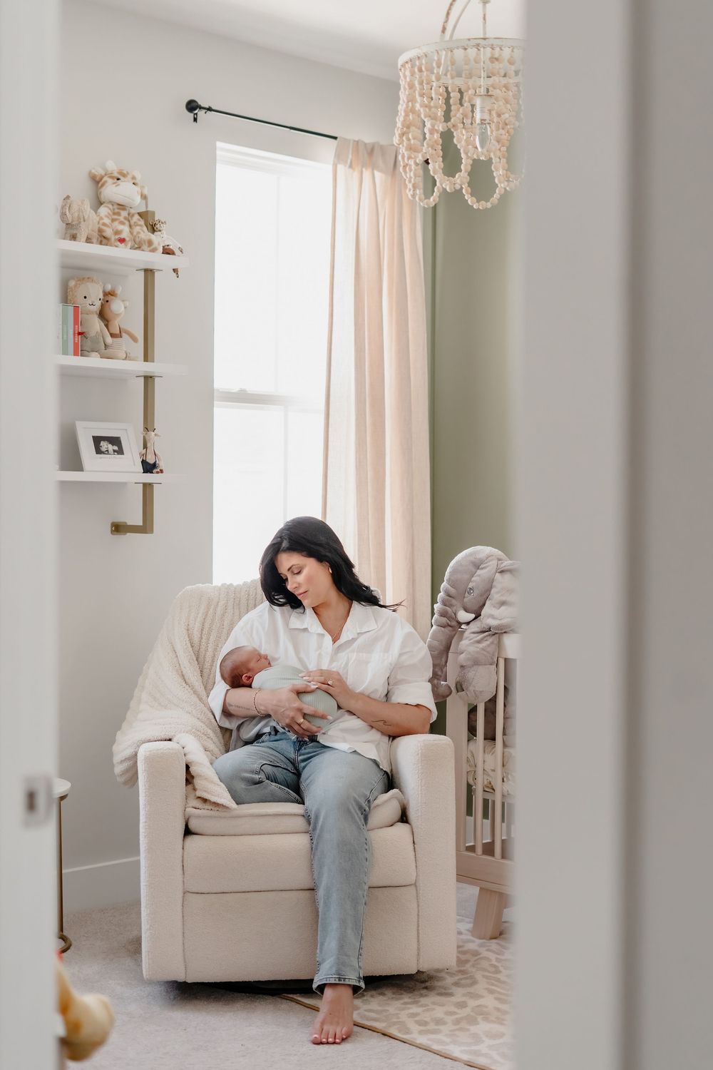 A parent in casual clothing sits on a white armchair in a bright, minimalist nursery feeding their newborn baby.