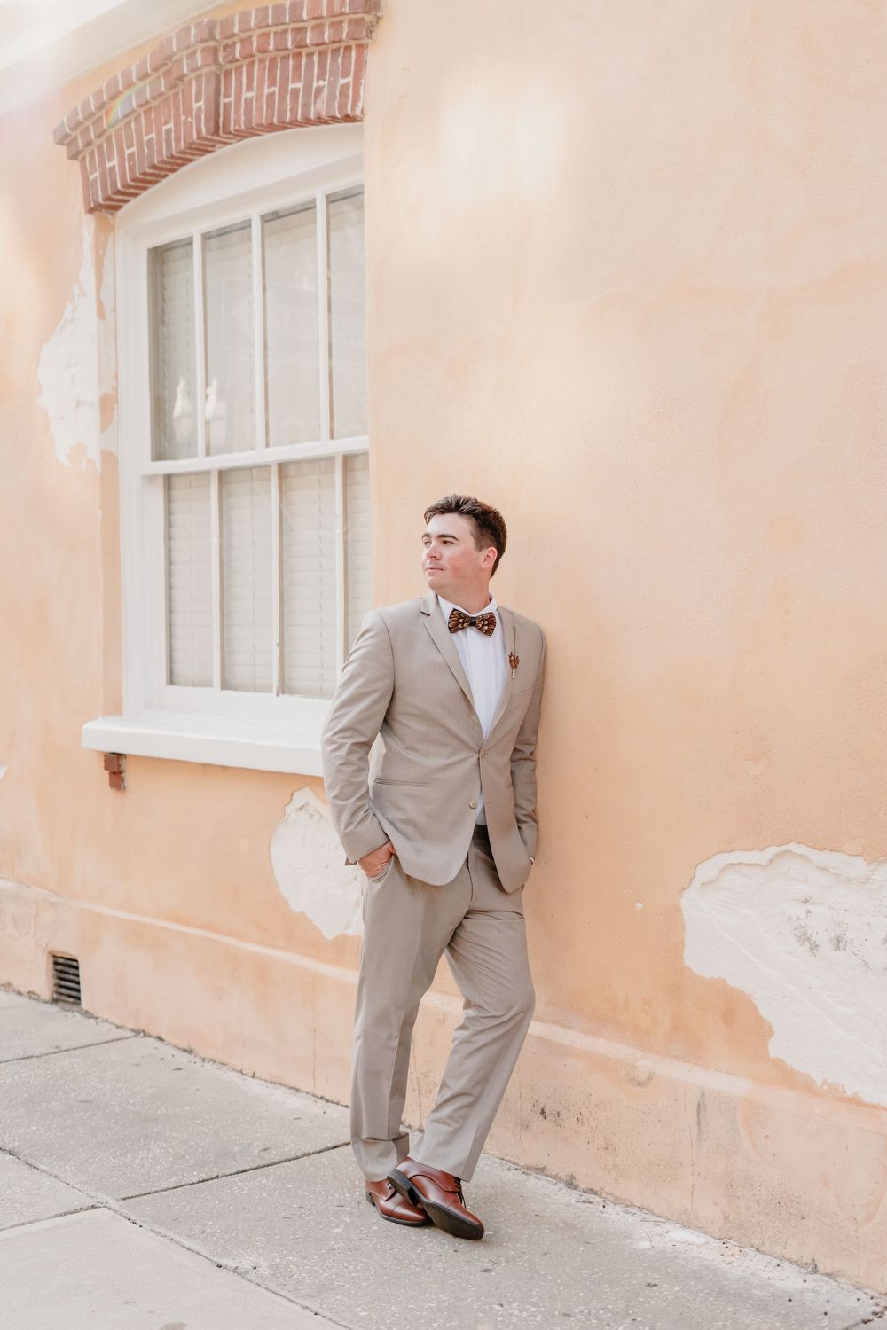 Stylishly dressed person in light beige suit and bow tie leaning against peachy colored wall with window in Charleston.
