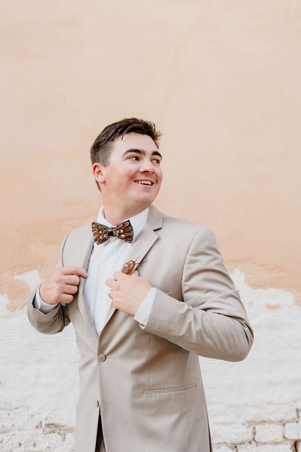 A person in a beige suit and leopard print bowtie poses happily in front of a light colored wall.