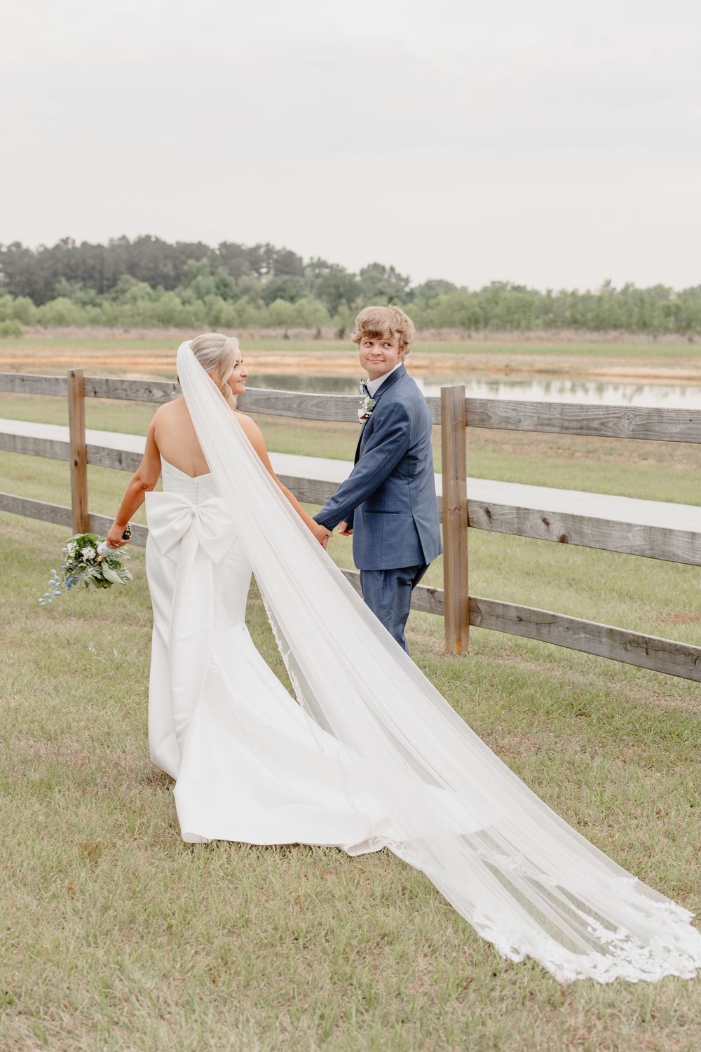 Bride and groom walk hand-in-hand along a wooden fence in a green field, bride wearing a long veil and bow-back gown.