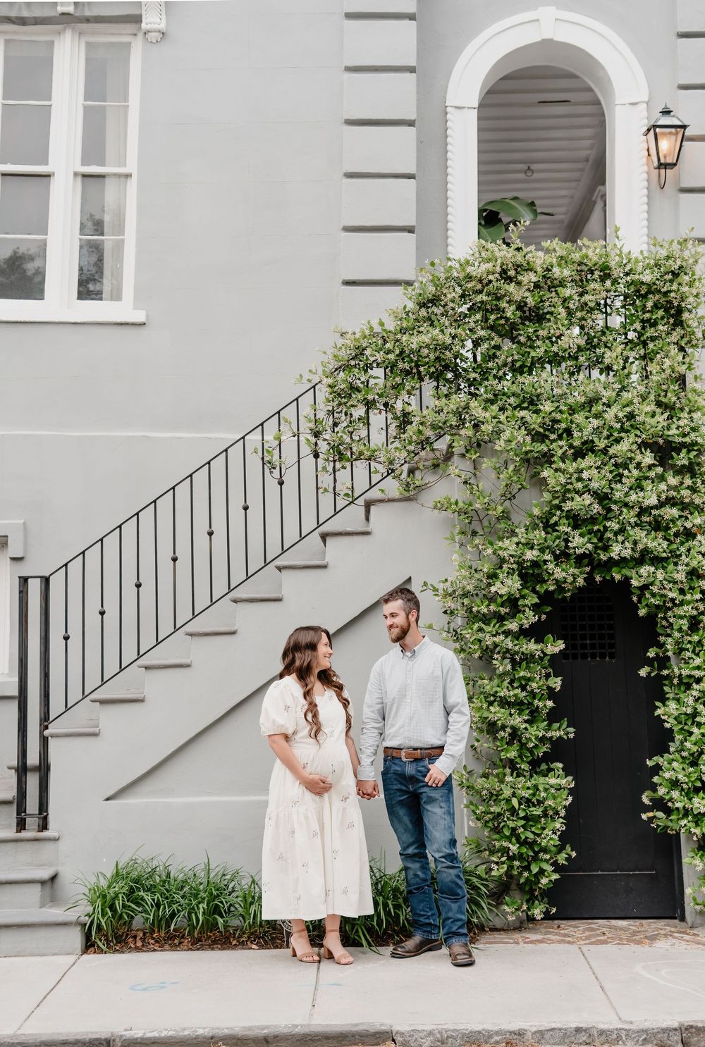 Couple stands together by an elegant white staircase with wrought iron railings and climbing vines in Charleston.