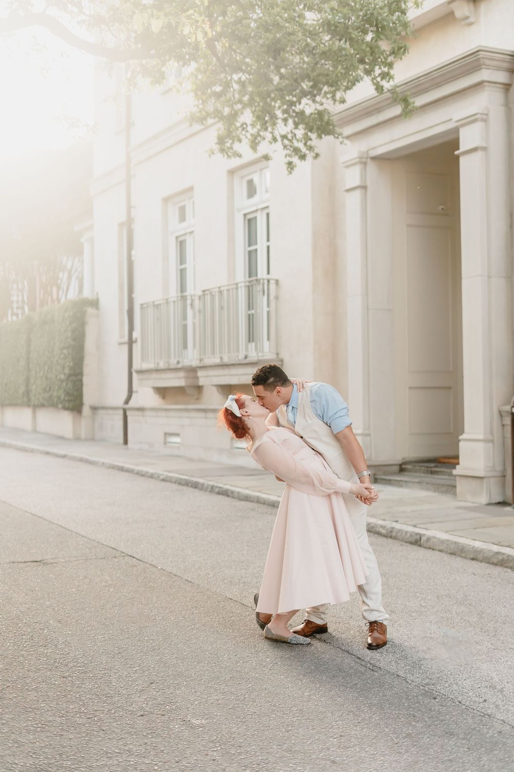 A romantic couple in vintage-style clothing share a tender dip kiss on a quiet street in front of a classic white building.