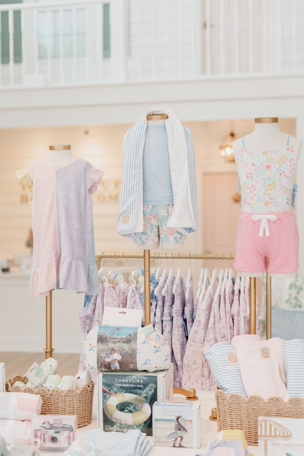Pastel baby clothing displayed in boutique with floral prints, ruffles, and wicker baskets on wooden shelves.