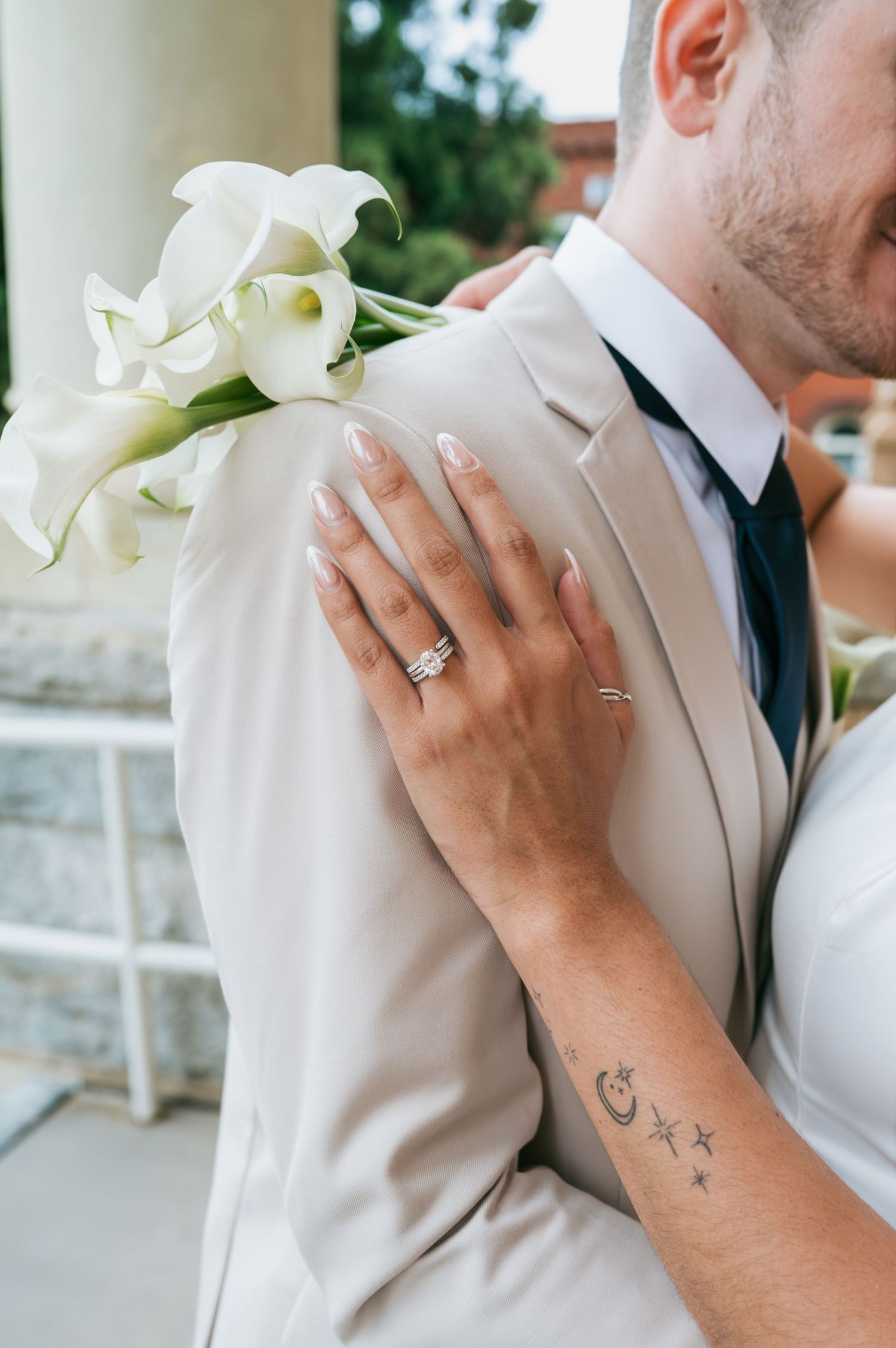 Close-up of bride's hand with engagement ring resting on groom's beige suit holding calla lilies Asheboro NC