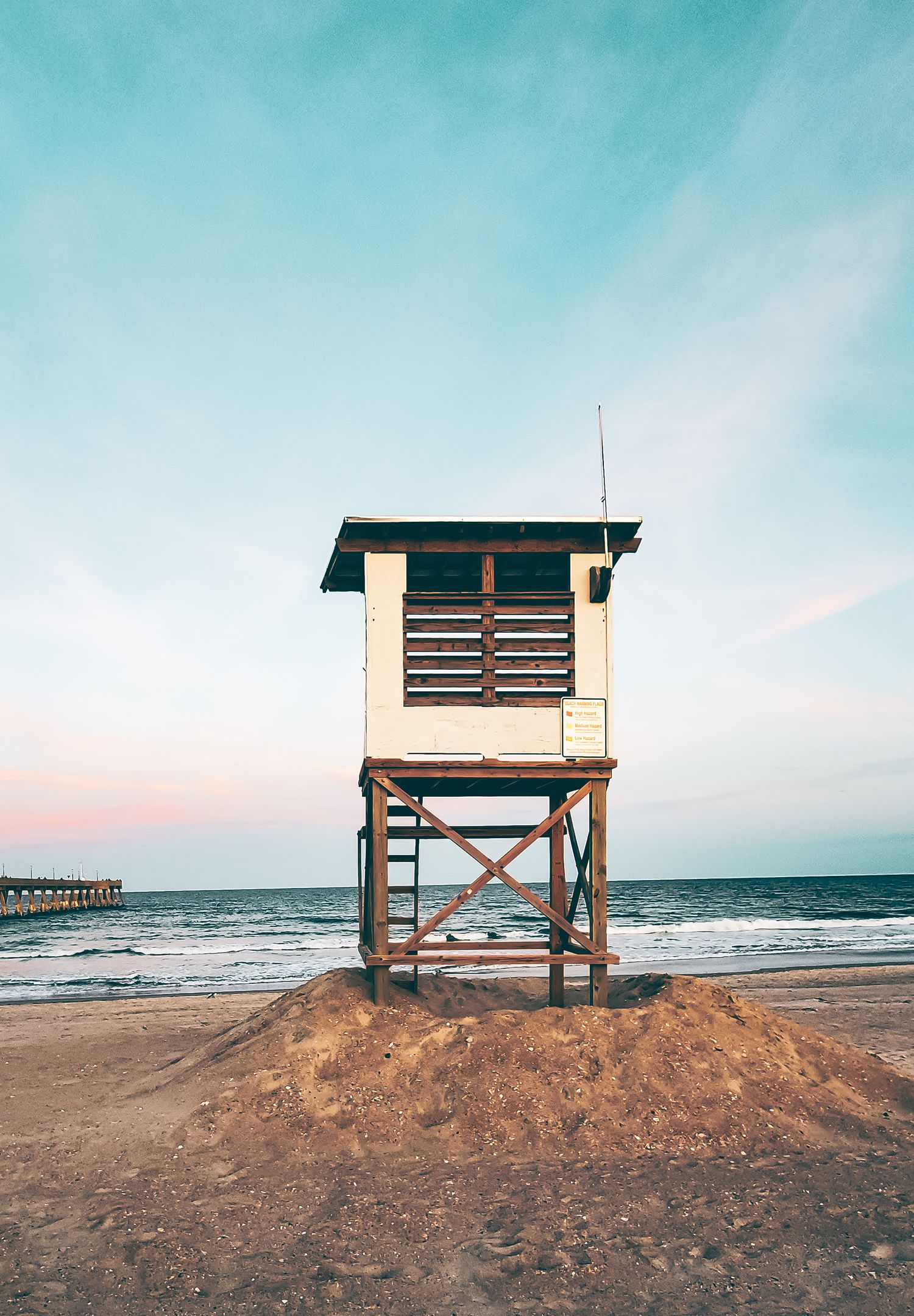 Wooden lifeguard tower stands on sandy beach at sunset with ocean pier in background.