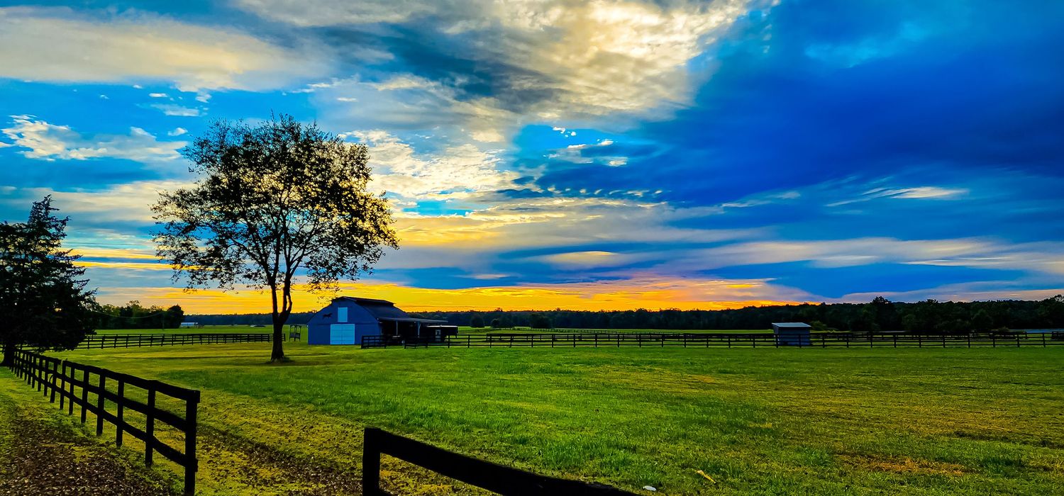 Rural farmland landscape with blue barn and fence under dramatic sunset sky.